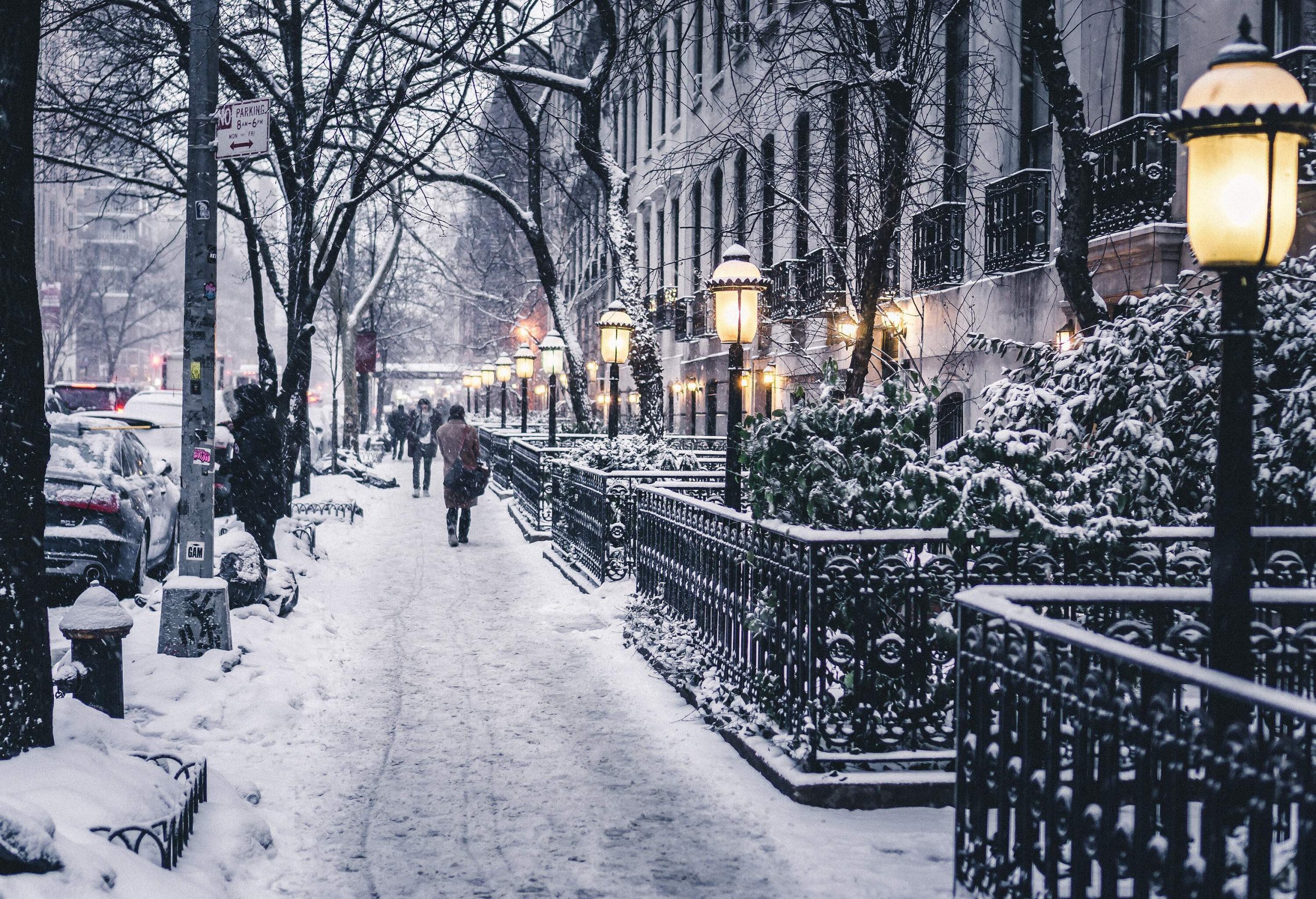 People strolling on a snow-covered sidewalk between parked cars and nearby buildings, bordered by barren trees and lit lamp posts.