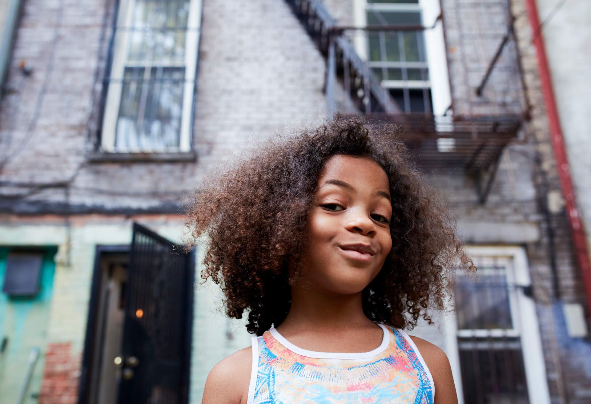 A little girl with curly hair standing in front of a tenement structure.