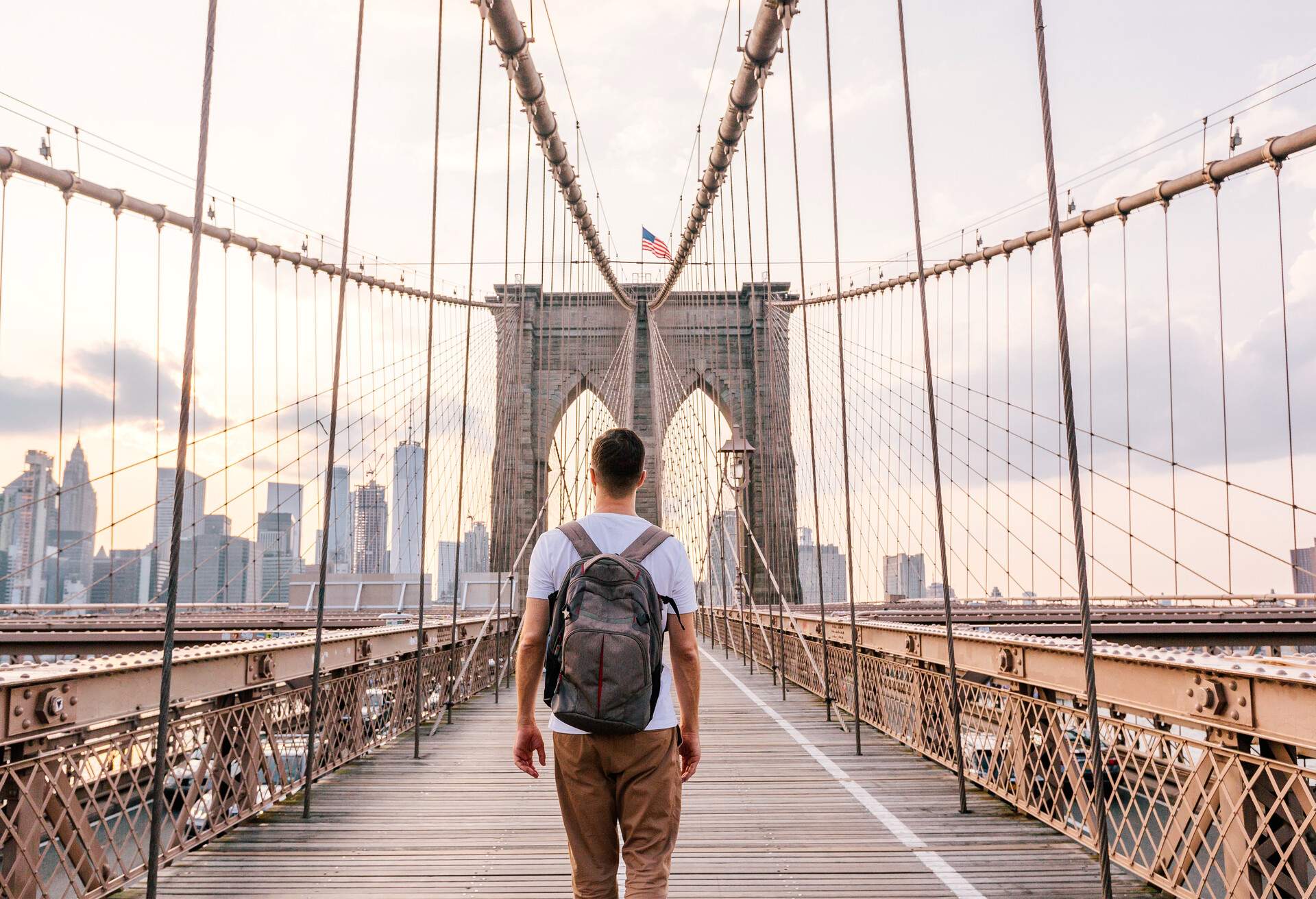 A man with a backpack walks on a footbridge with suspension cables against the backdrop of the city skyline.