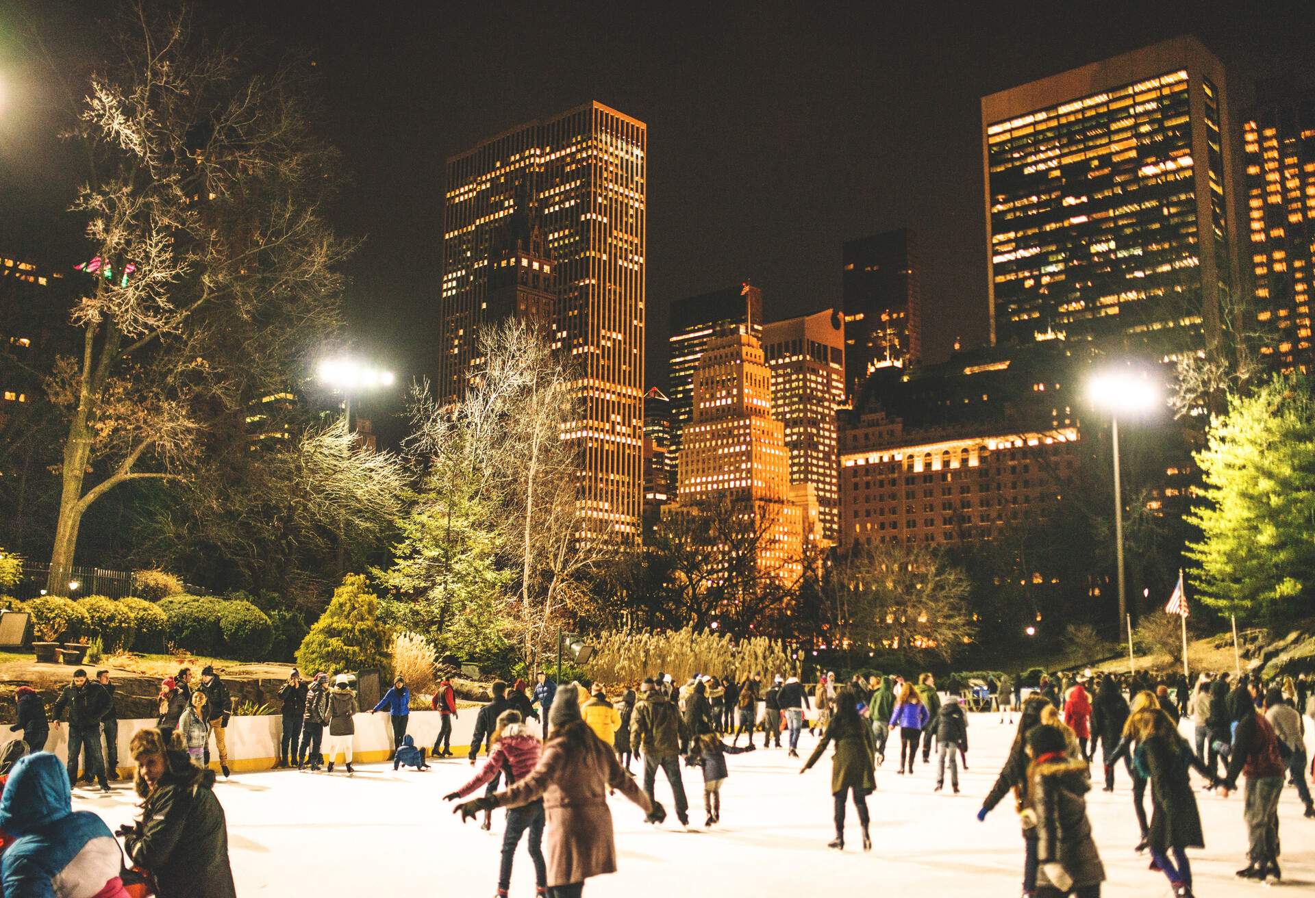 People ice skating in Central Park surrounded by tall trees with the illuminated skyscrapers of Midtown New York City in the background.