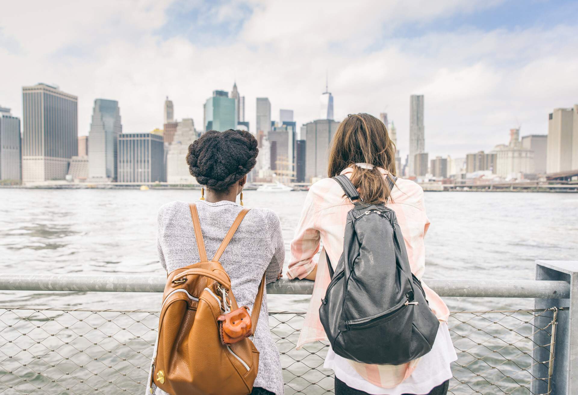 Two females leaning on a railing by the lake as they take in the view of the New York skyline.