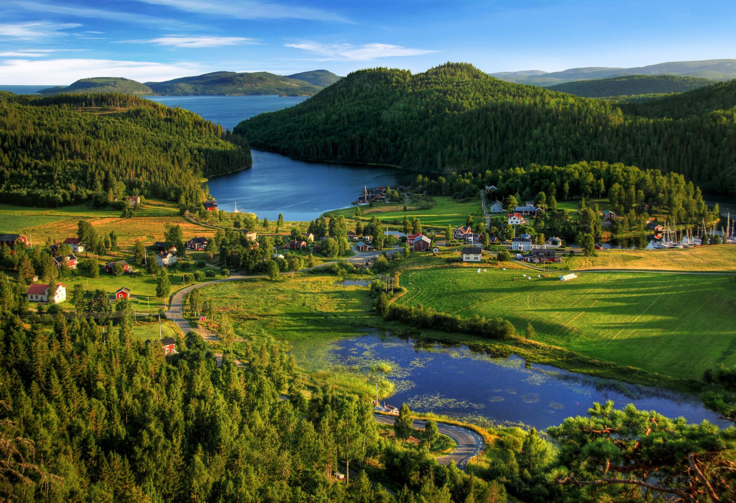 A road along a pond that leads to a village beside a lake and forested hills.