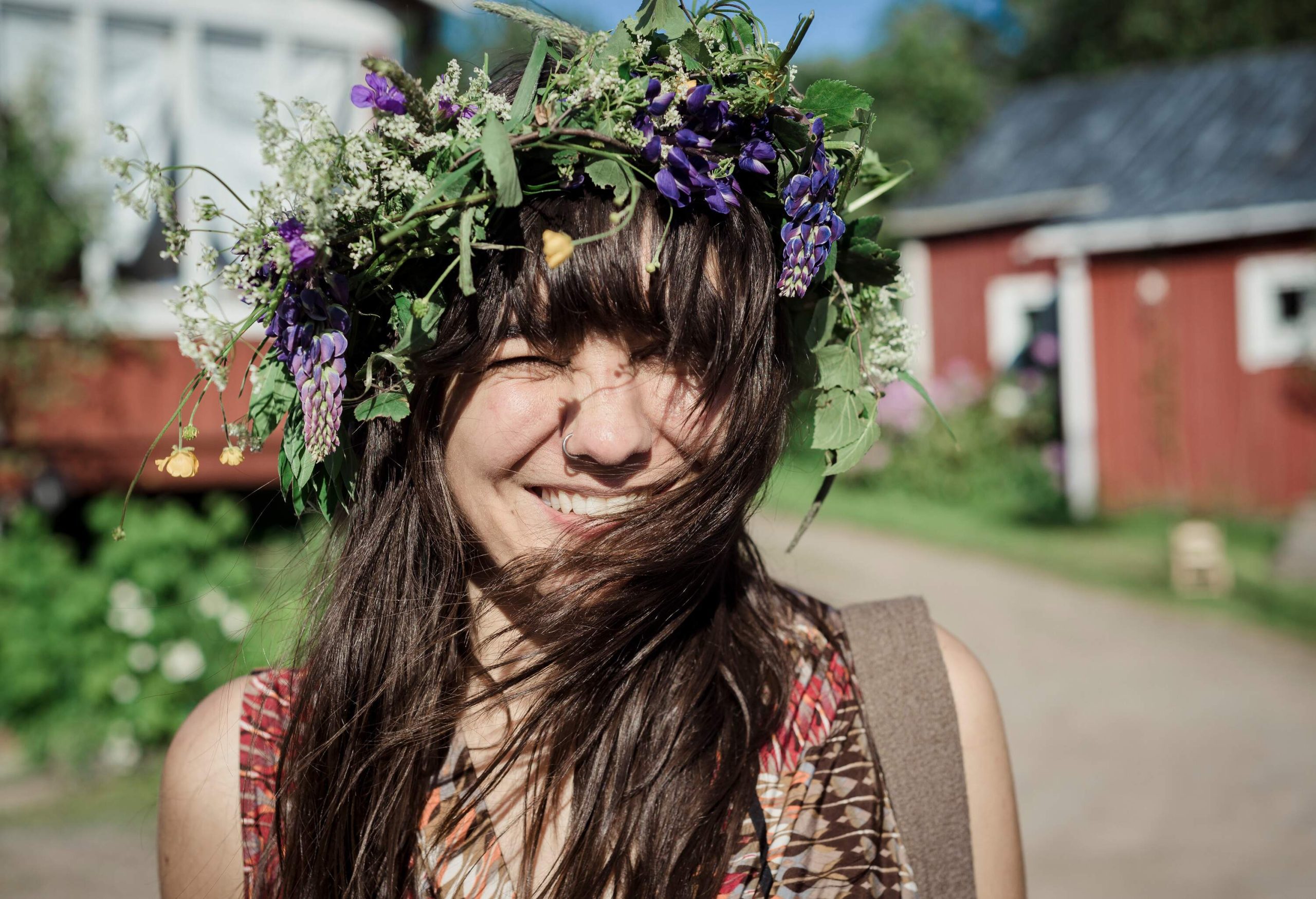 A long-haired, smiling young lady wears a floral wreath with her hair blown by the wind covering her chin.
