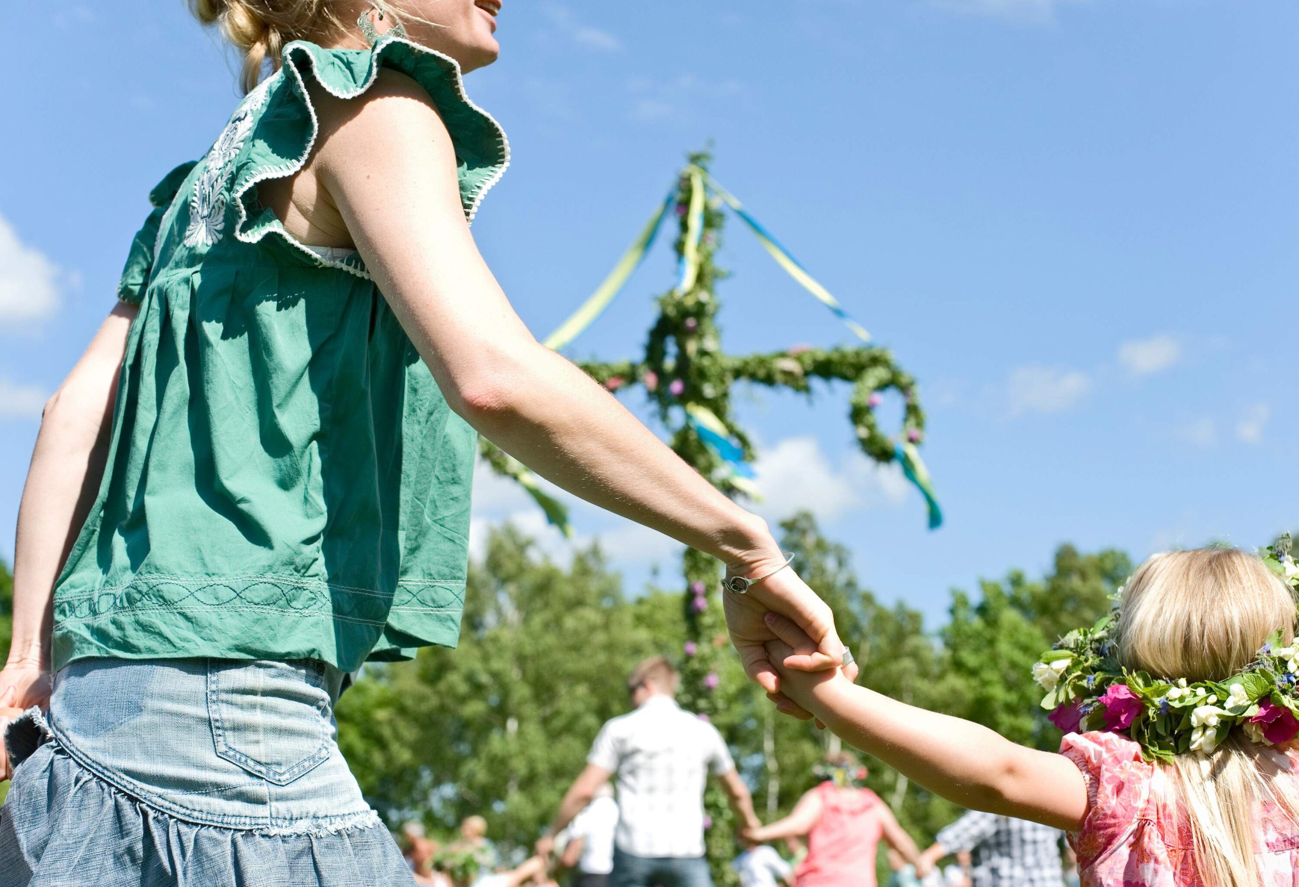 A mother holds her daughter's hand, wearing a floral wreath near a maypole with ribbons covered with flowers and leaves.