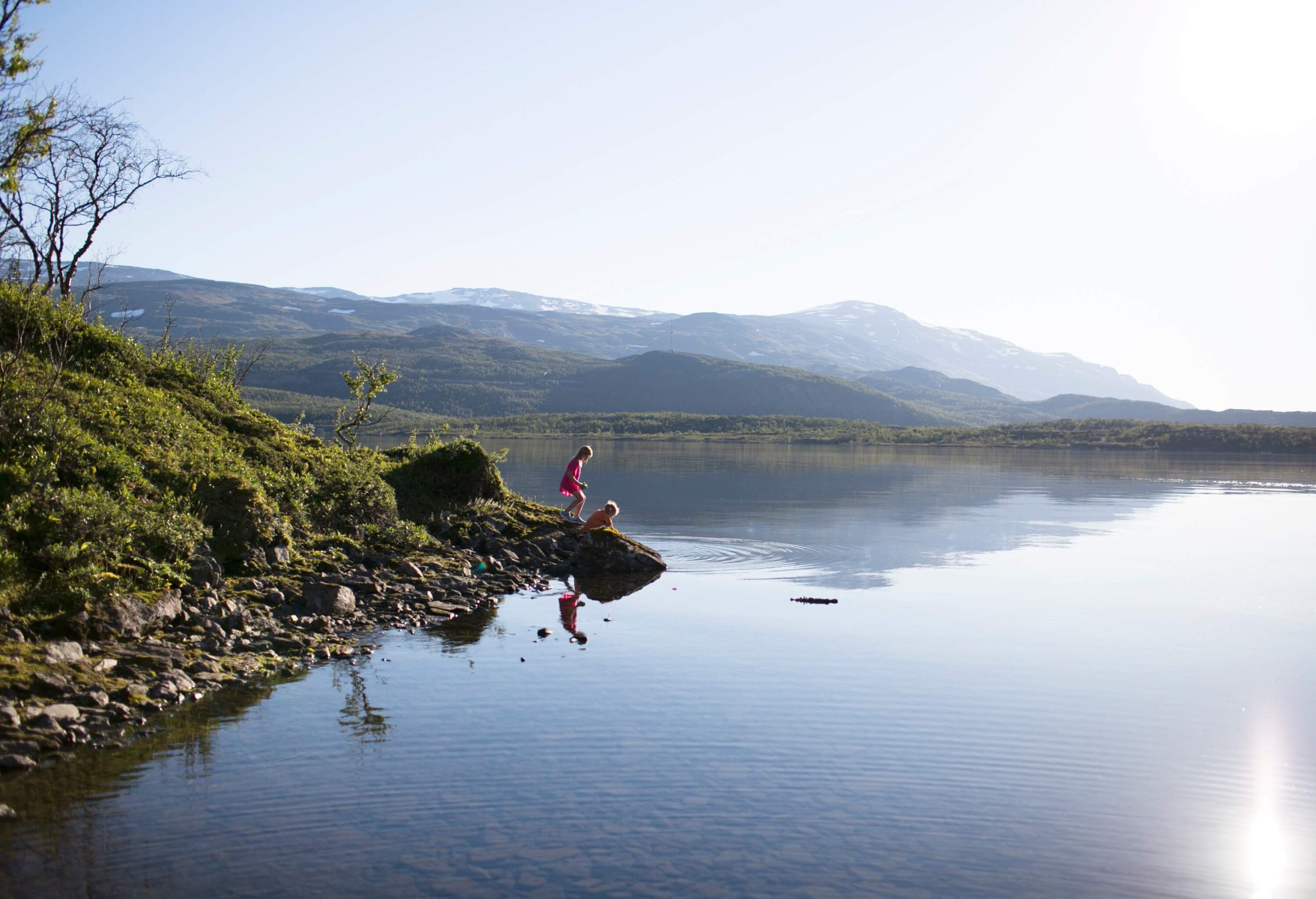 Two kids playing in the water while standing on a rock on the shore of a lake.