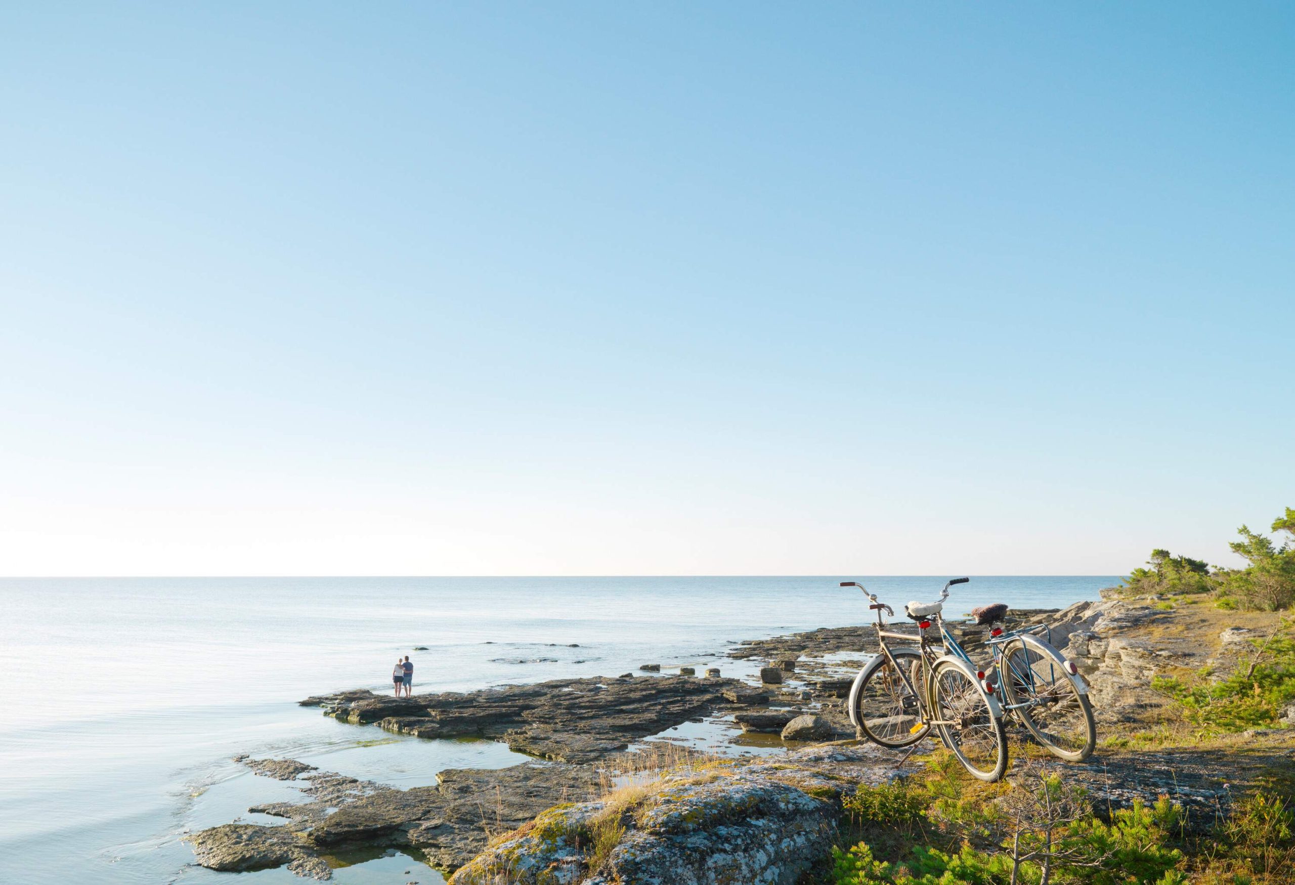 A couple stands on a rocky beach gazing out to sea with their bicycles parked in the foreground.