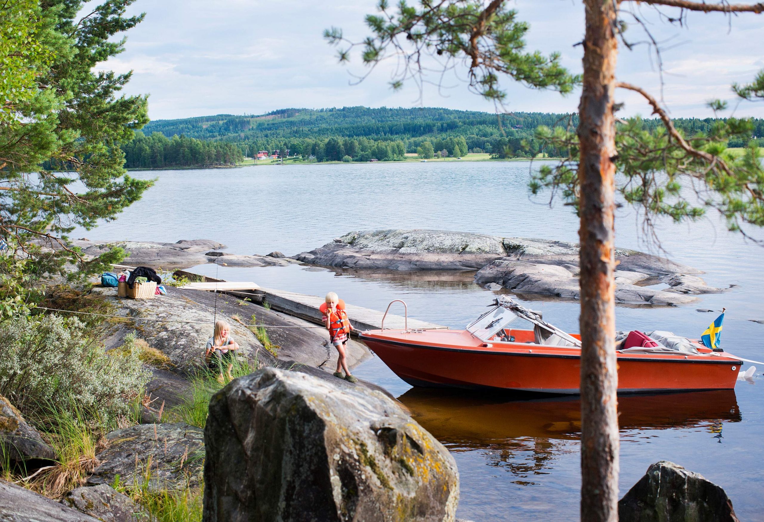 View of a tree by a lake surrounded by rock, an orange boat and a child coming out of the water on a cloudy summer day