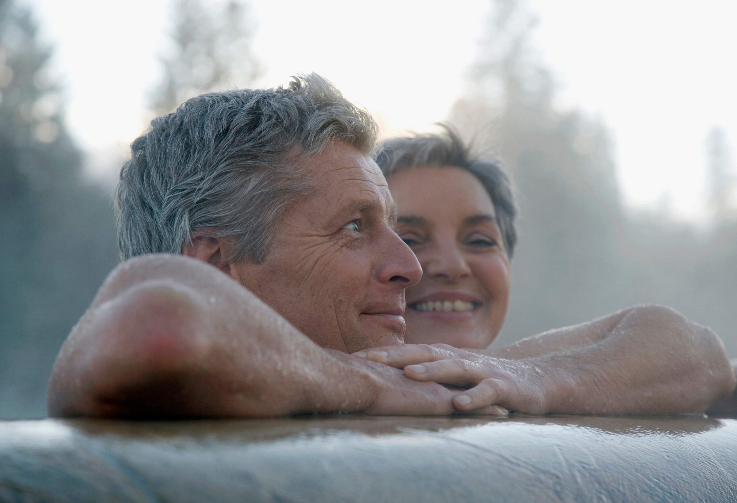 An elderly couple smiles while leaning on the edge of a jacuzzi.