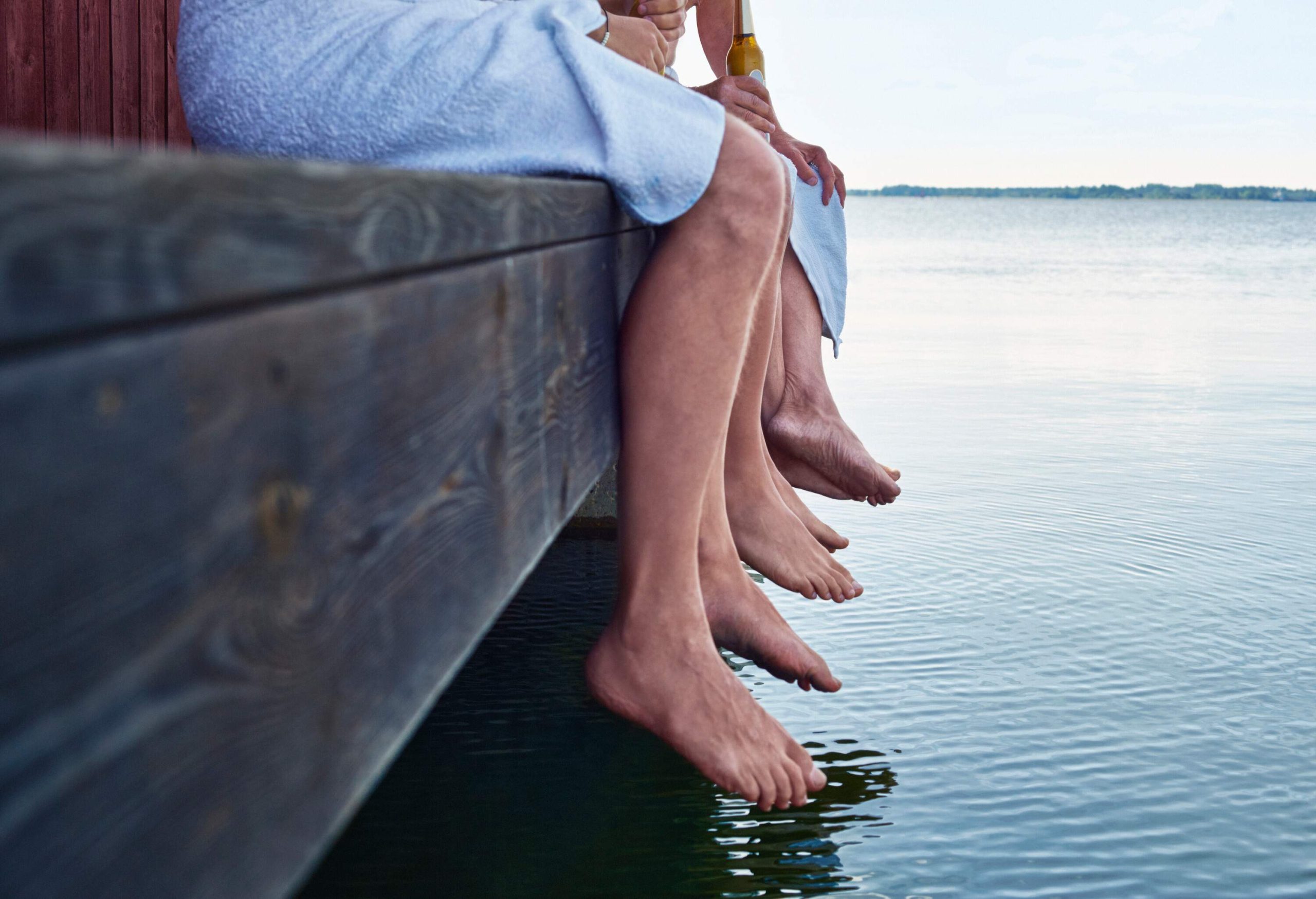 Three pairs of legs hanging on a wooden platform above a lake.