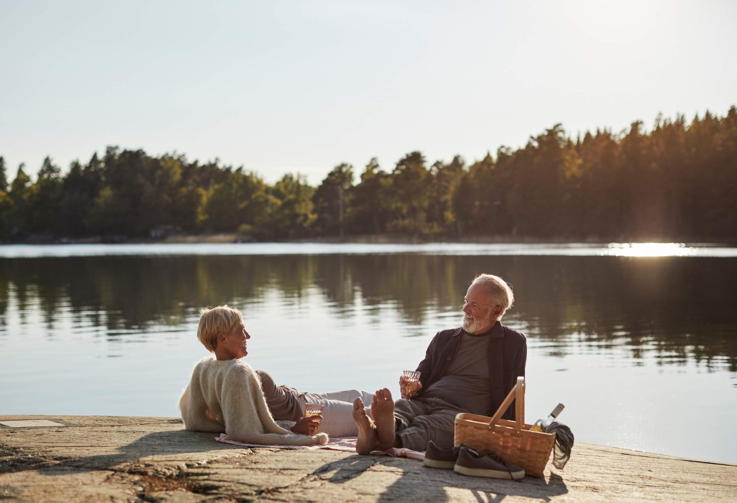 An elderly couple hanging out with a glass of drinks alongside a tranquil lake surrounded by trees.