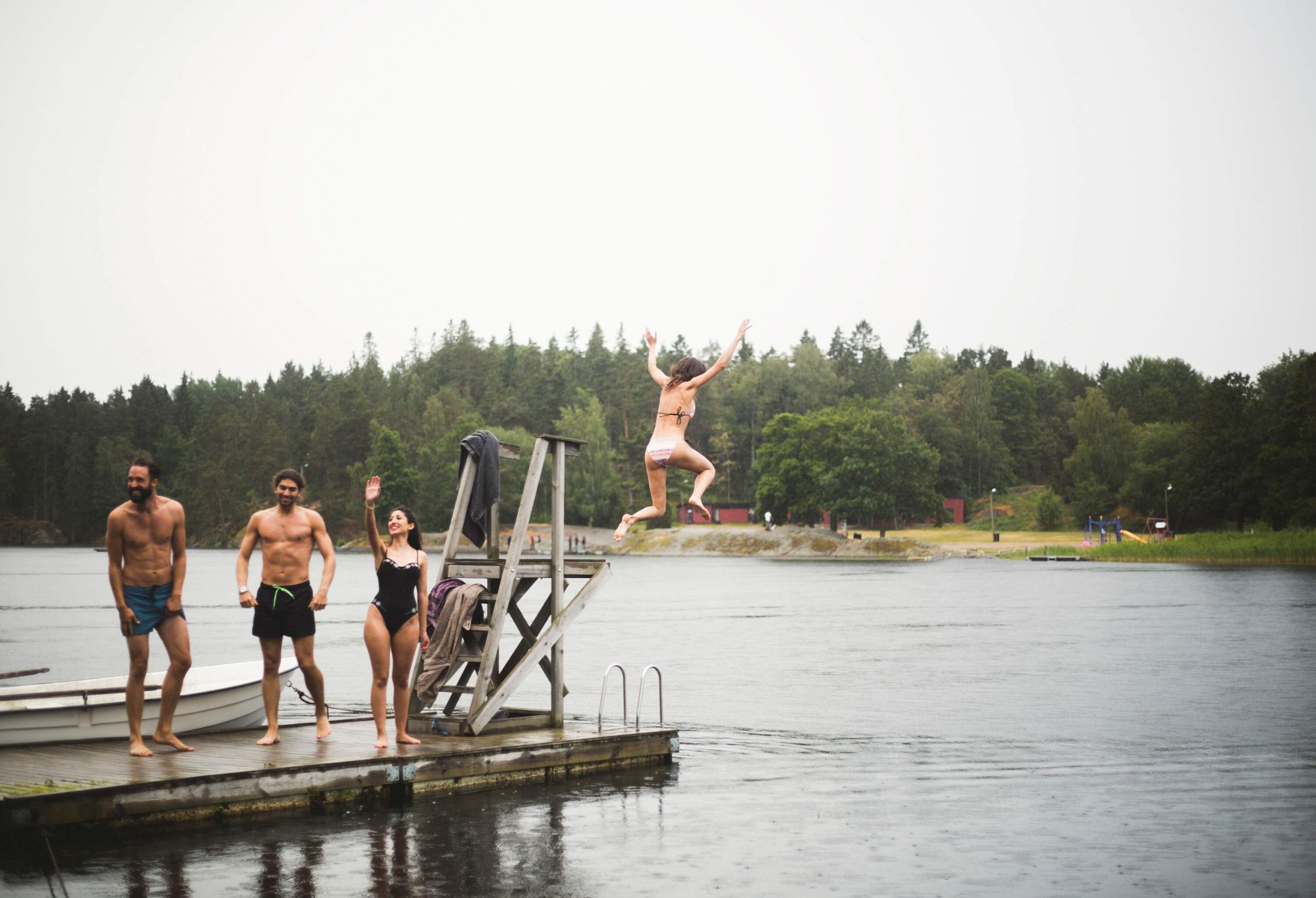 Three people in swimsuits stand on a wooden jetty as a woman leaps from a ledge into a lake.