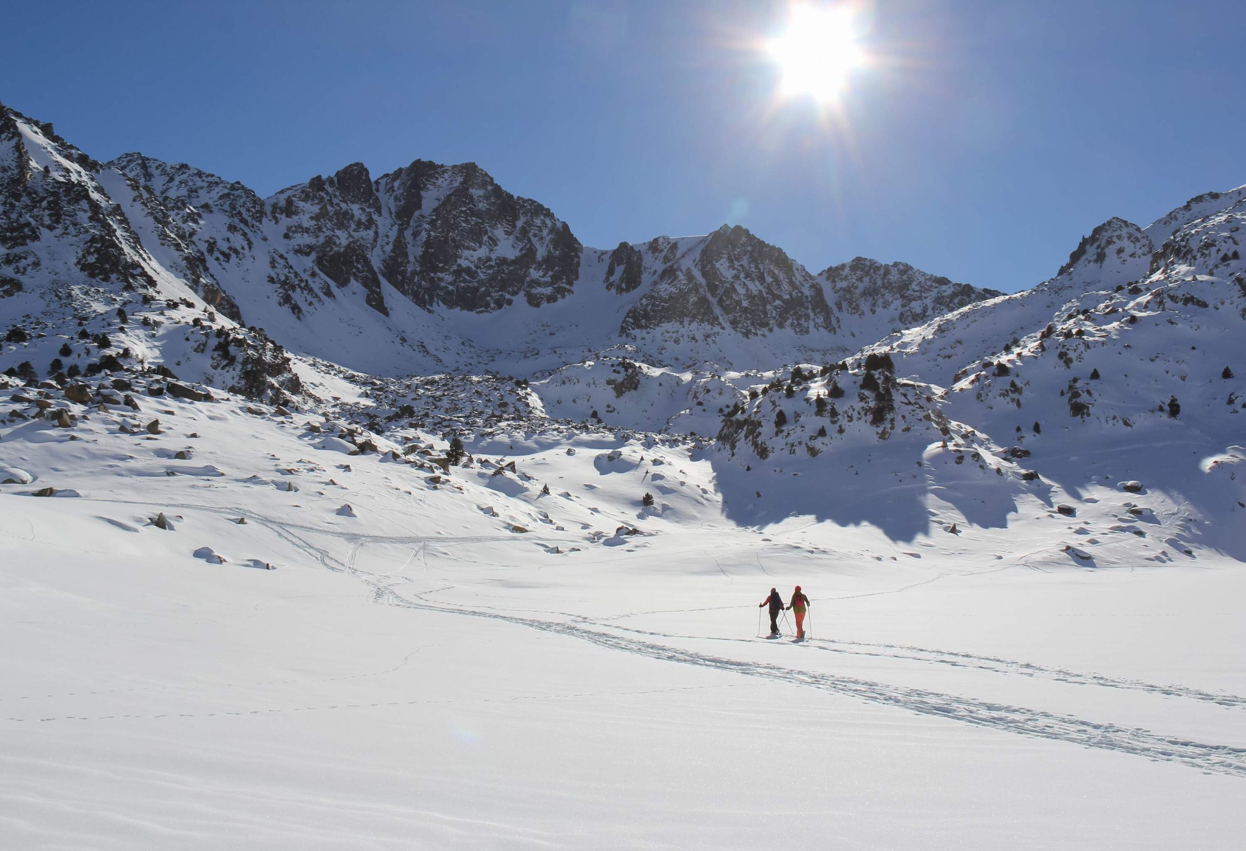 Two skiers walk through powder snow land beneath the mountains covered in deep snow.