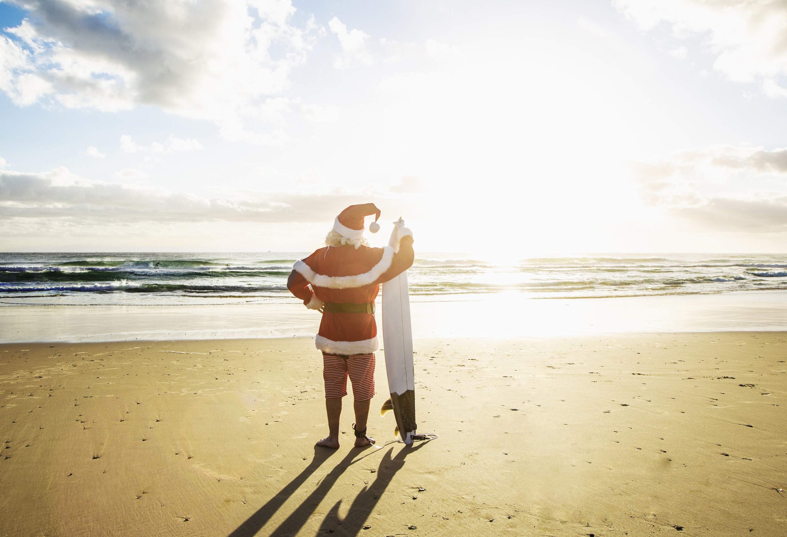 Santa Claus standing on the sand of the beach facing the water while holding a surfboard.