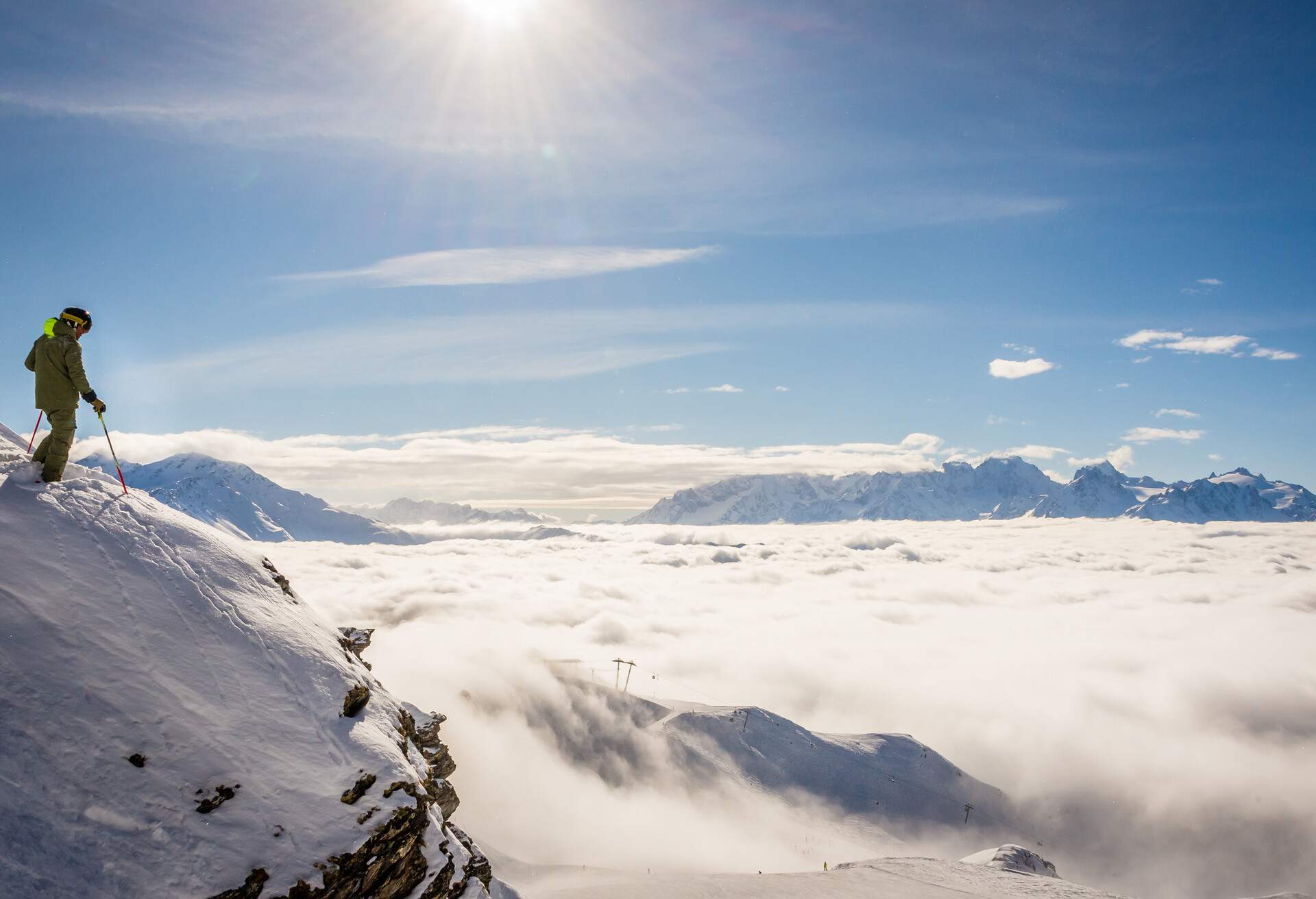 A skier atop a cliff looking down on an overcast layer of clouds across the mountains in the Alps.
