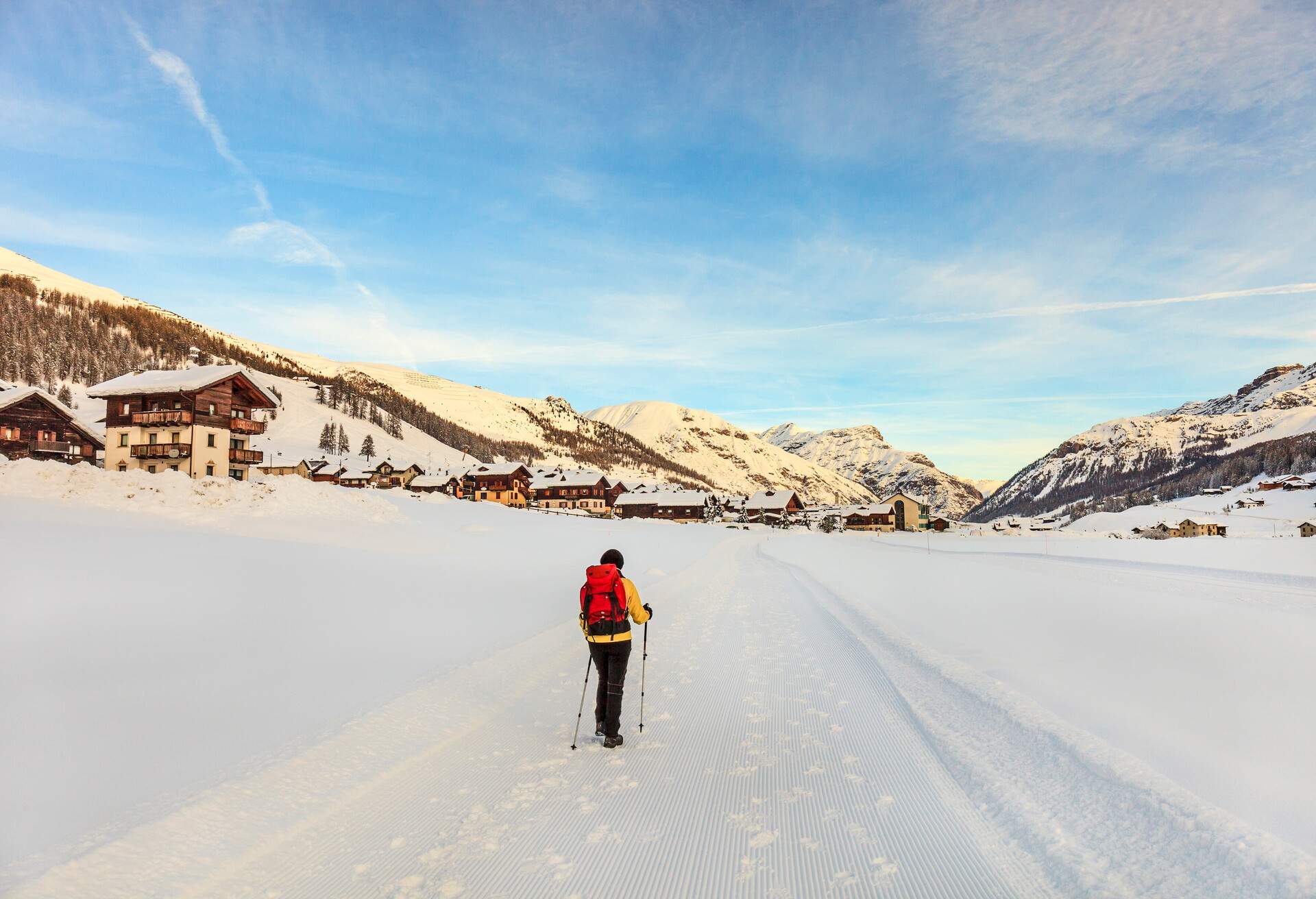 A woman wearing yellow winter clothes and holding two trekking poles is walking on a snowy road overlooking a ski resort under a snowcapped mountain.