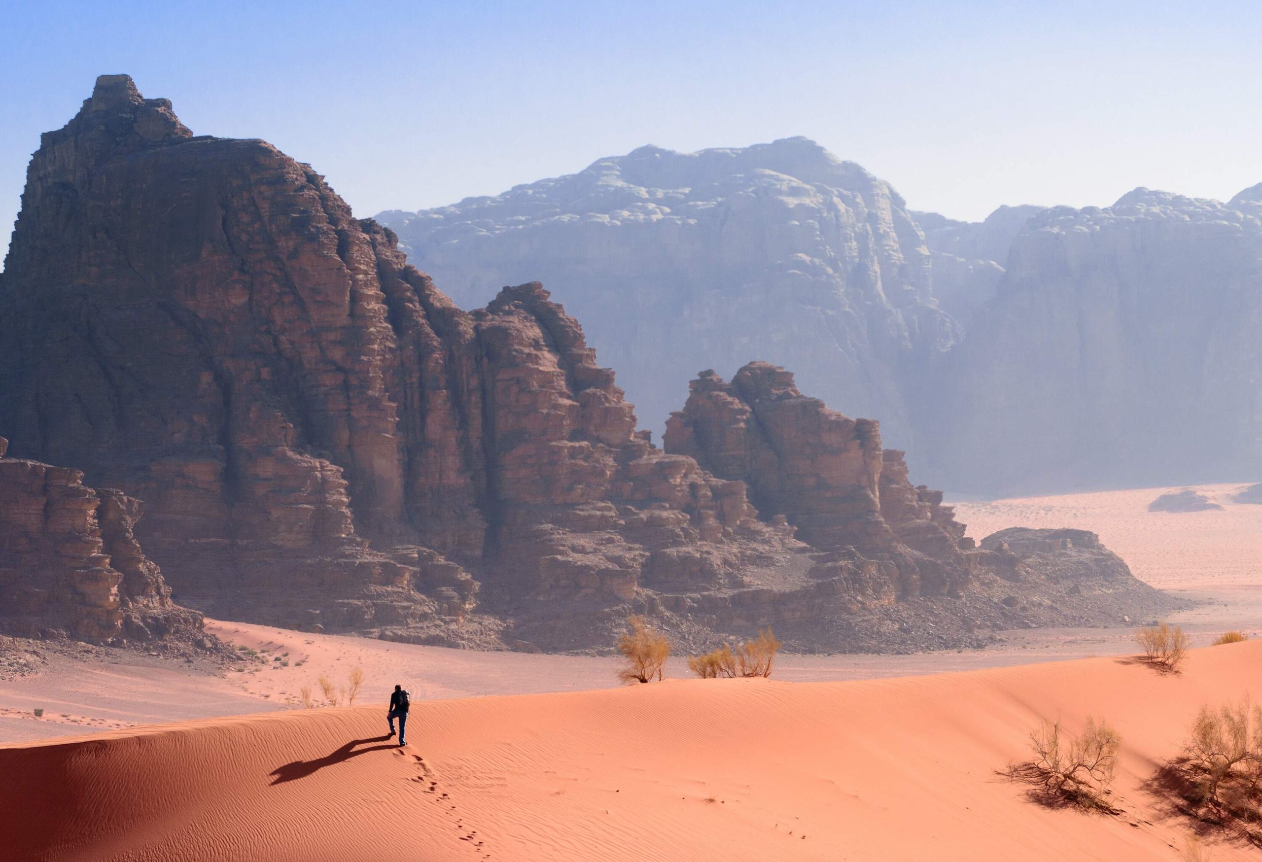 A man walking across a sand dune towards the sandstone mountains in the valley.
