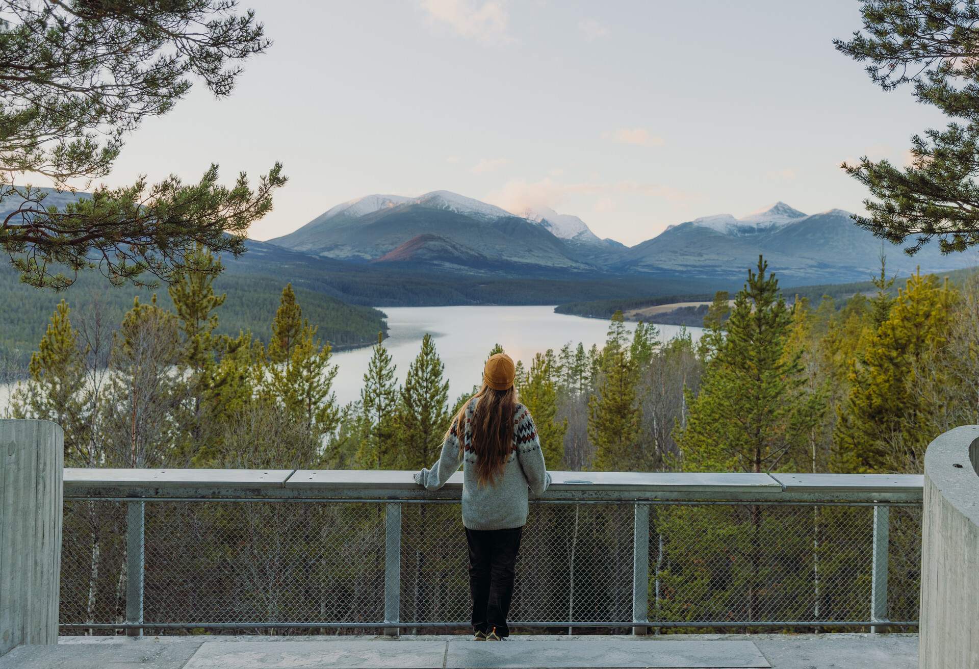 Rear view of female with long hair in yellow hat staying at the viewpoint admiring the scenic mountain landscape with lake and the pine forest during sunset in Rondane National Park