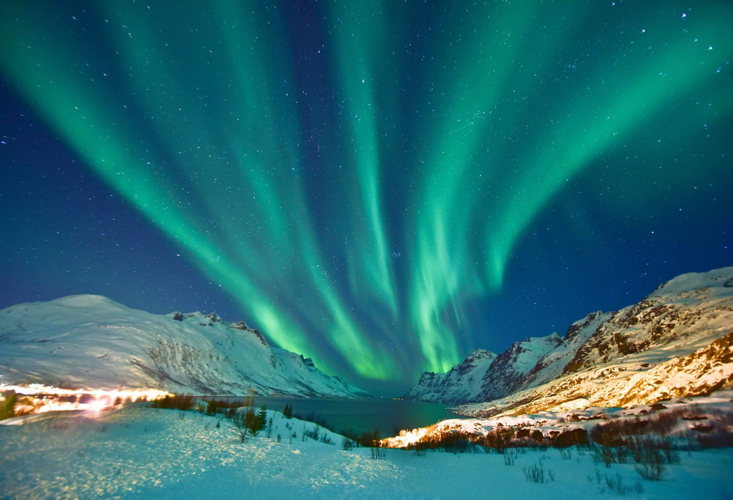 The majestic Aurora Borealis dances in the night sky above a snow-covered mountain landscape, casting a colourful reflection on a serene and frozen lake below.