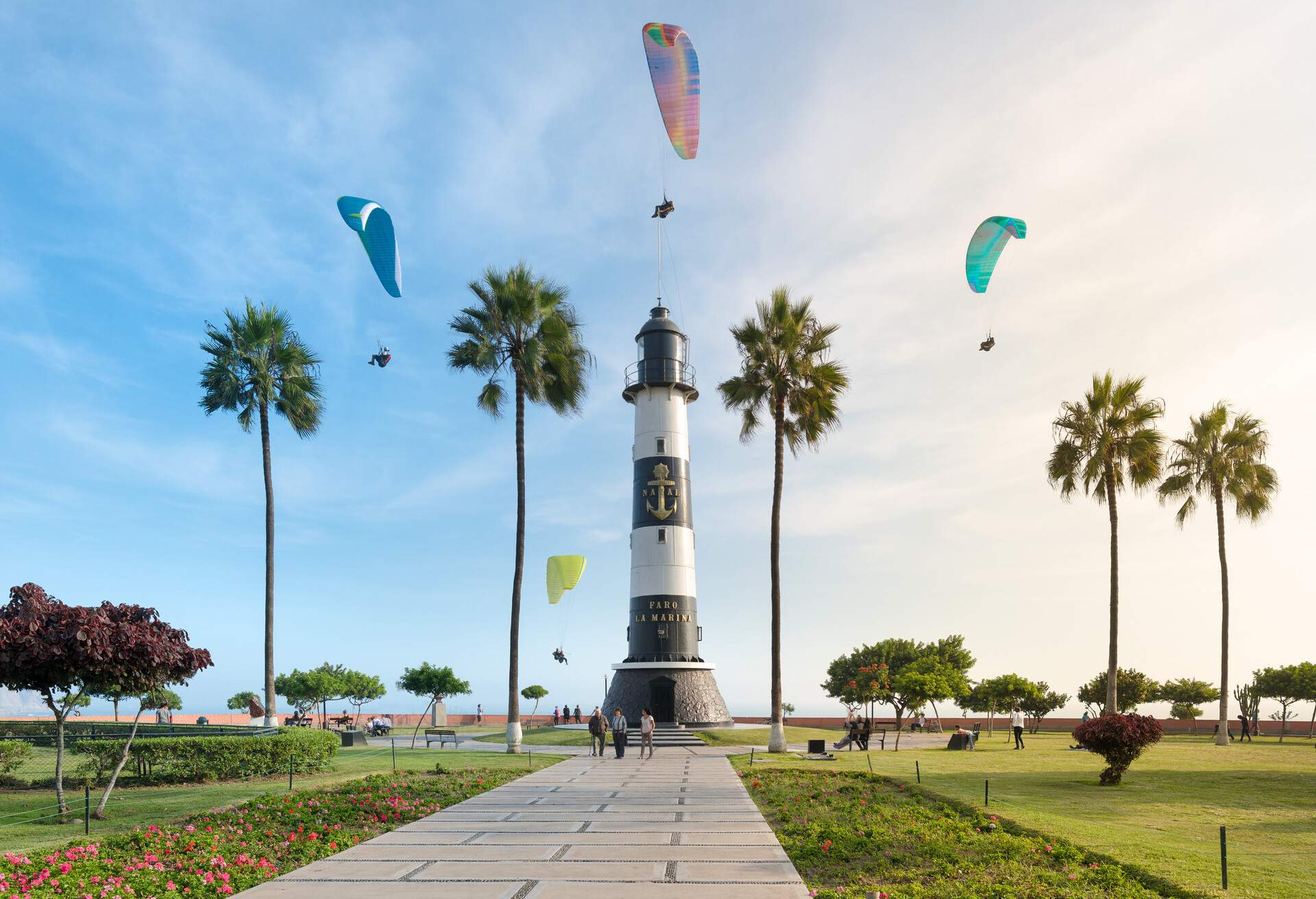 The Miraflores Lighthouse stands proudly at the heart of a coastal park, where towering palms dot the landscape, people meander along green turf, and paragliders gracefully soar above in the backdrop.
