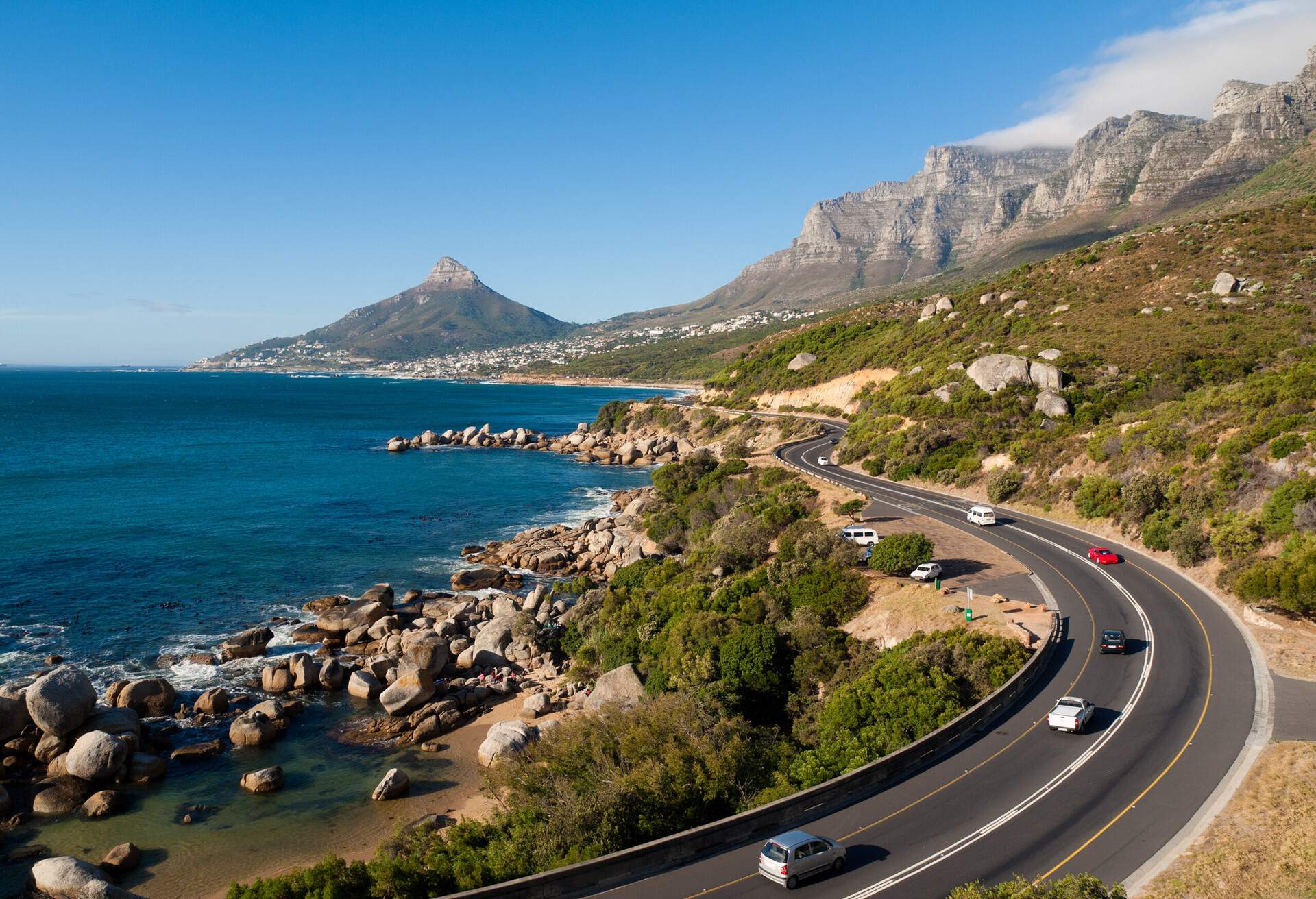 Cars travel across a coastal road along a forested slope with sweeping views of rugged mountains and a vast blue ocean.