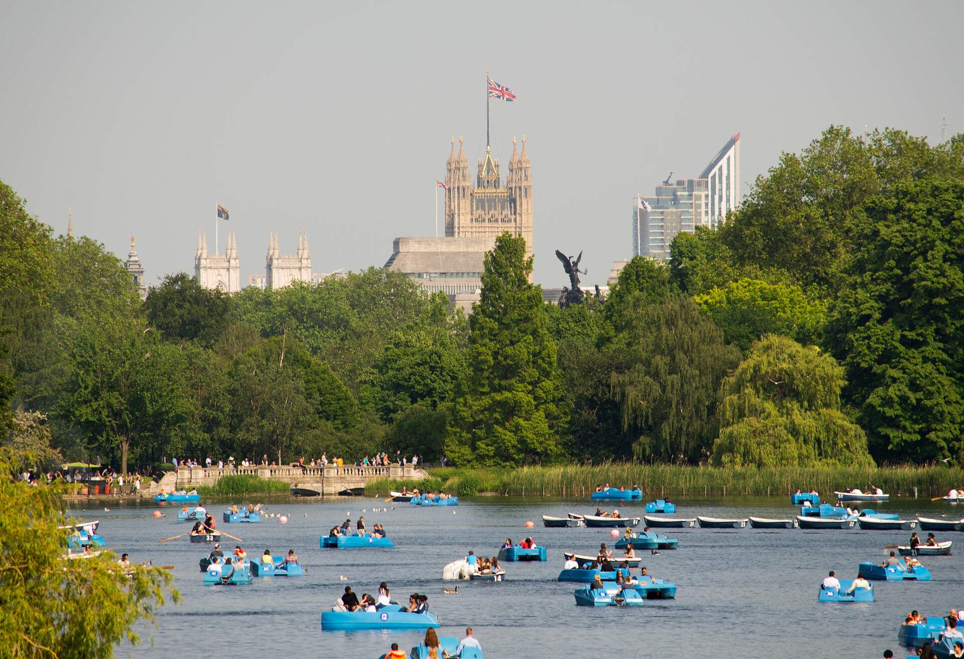 A crowd of people on blue boats in a lake lined with tall green trees with a view of the palace Westminster tower with a flag of United Kindom.