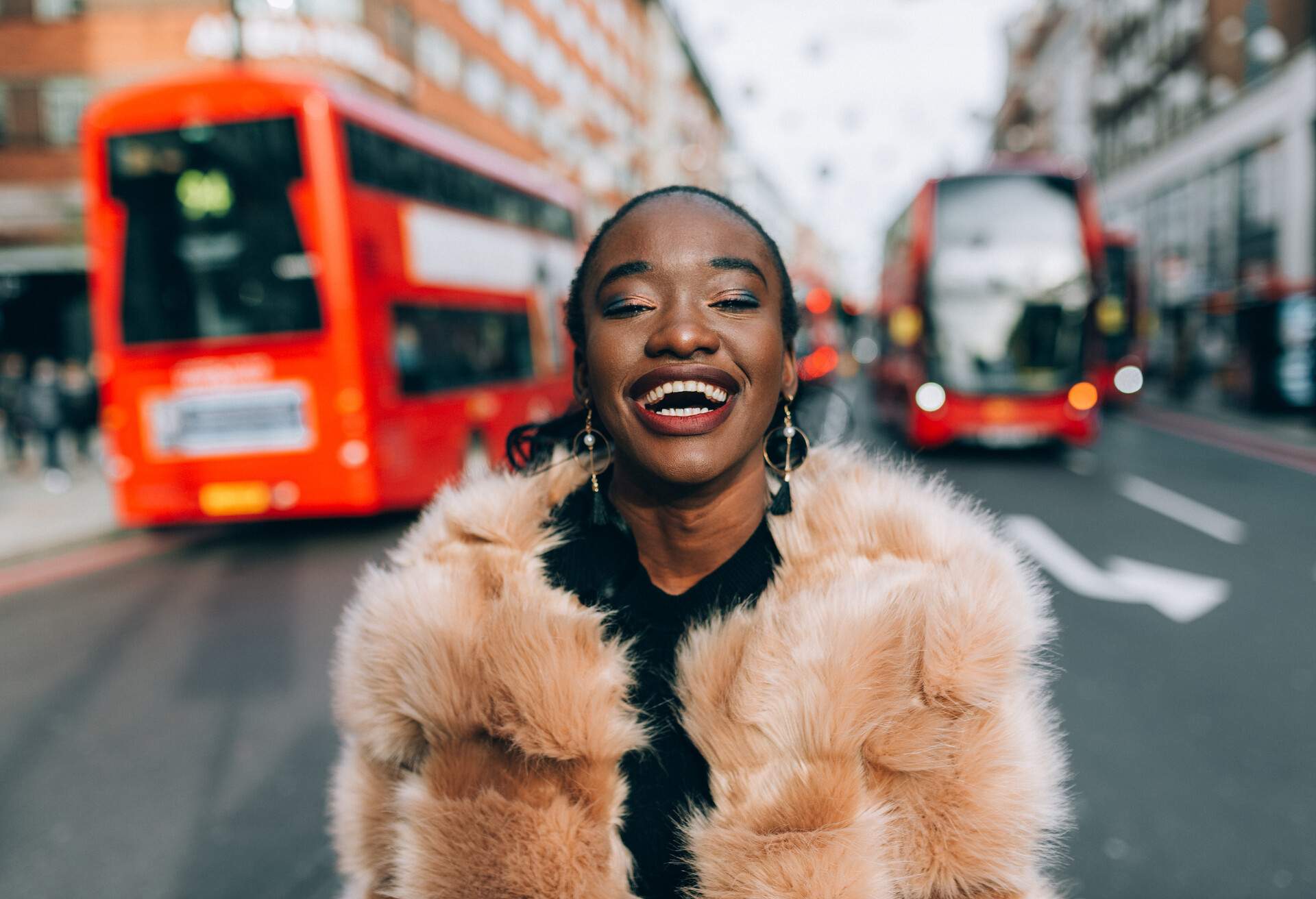 A happy dark-skinned lady with a fur jacket photographed in the middle of the street behind the red buses.