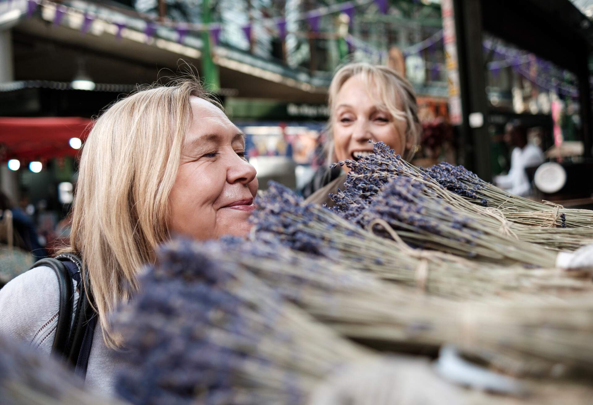 An older adult smells the pile of lavender while her companion watches her with a smile.