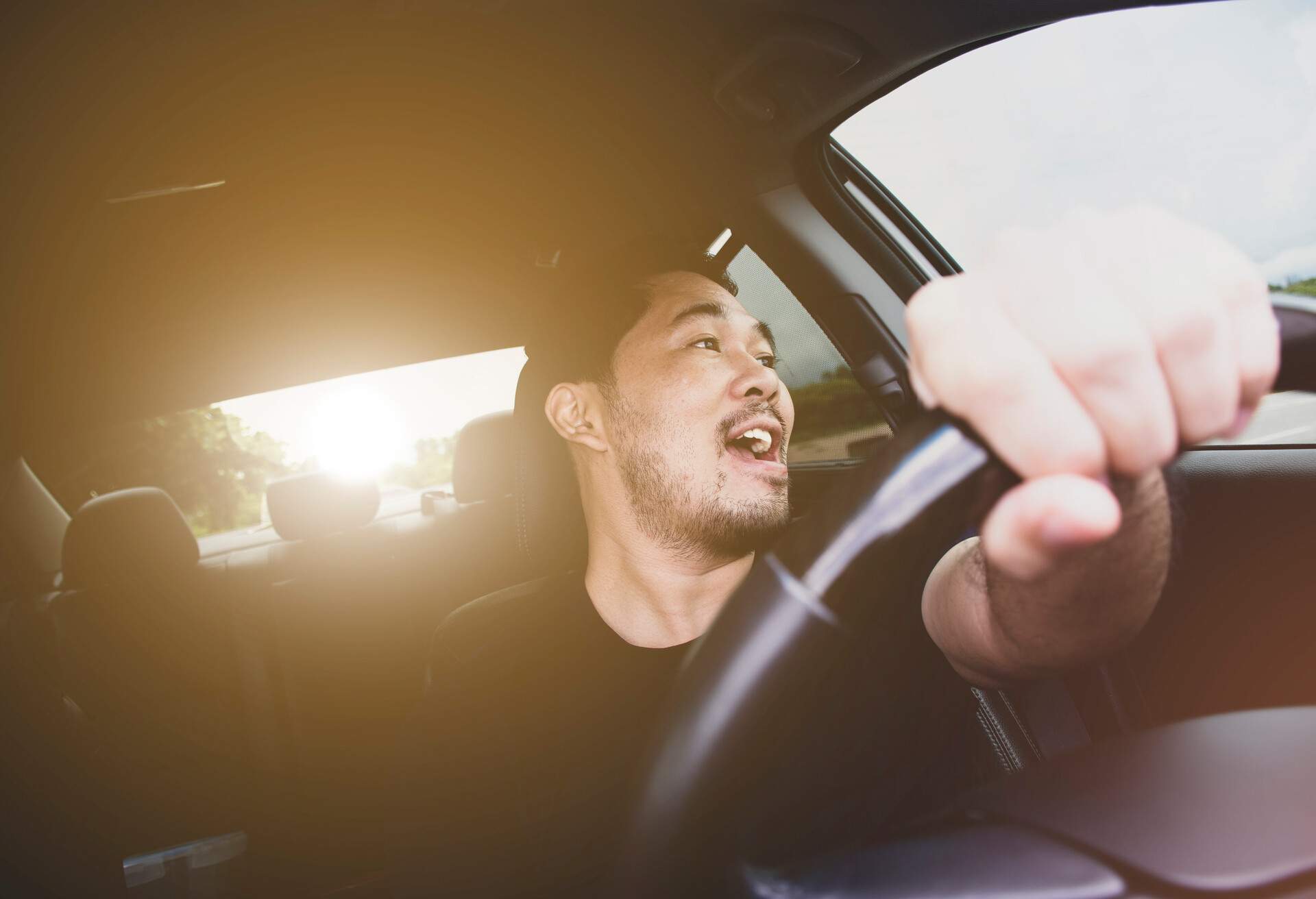 A man singing in the car while driving as the sun hits the rear windshield.