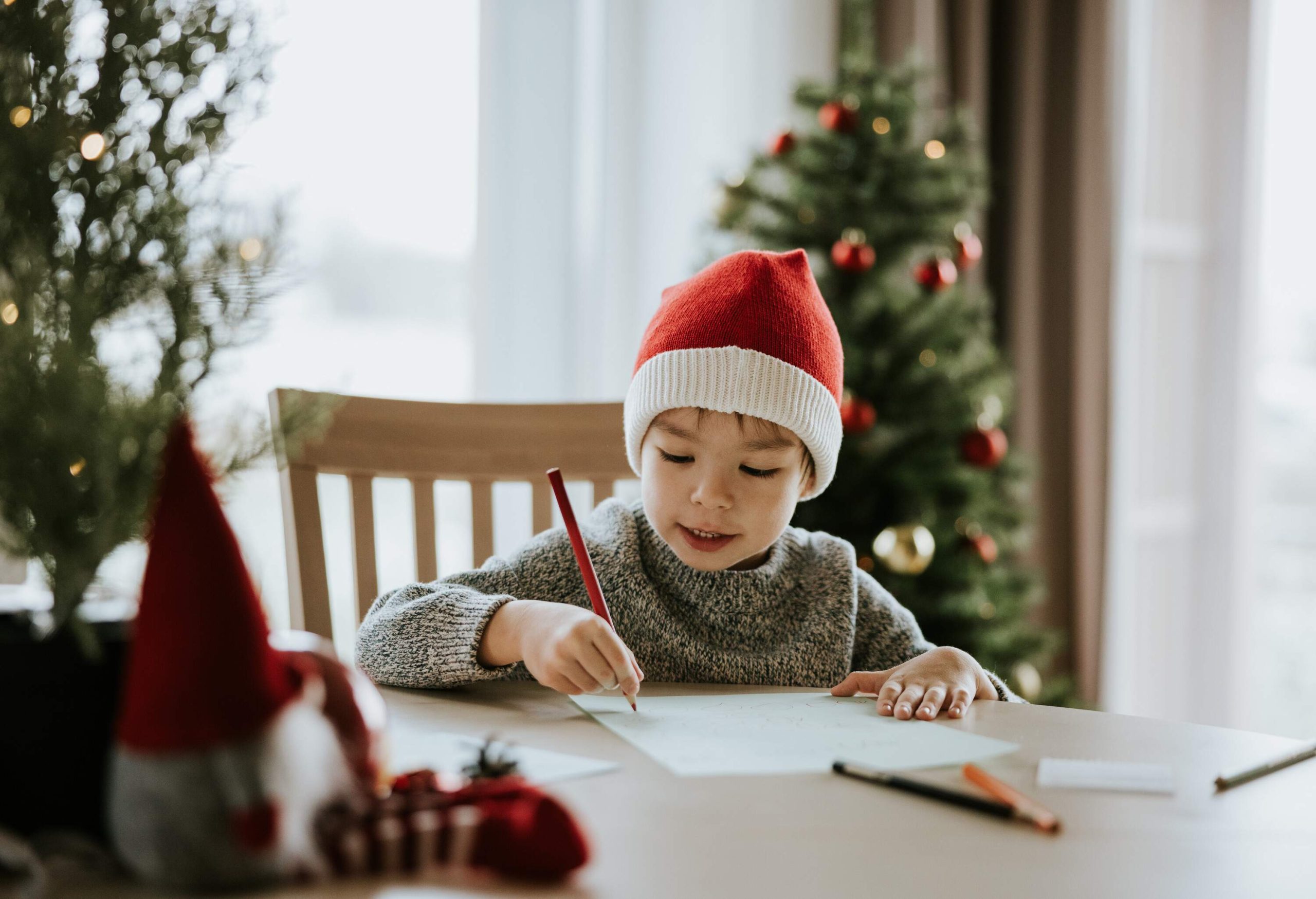 a little boy sitting by a table in a living room writing a letter while wearing a christmas hat