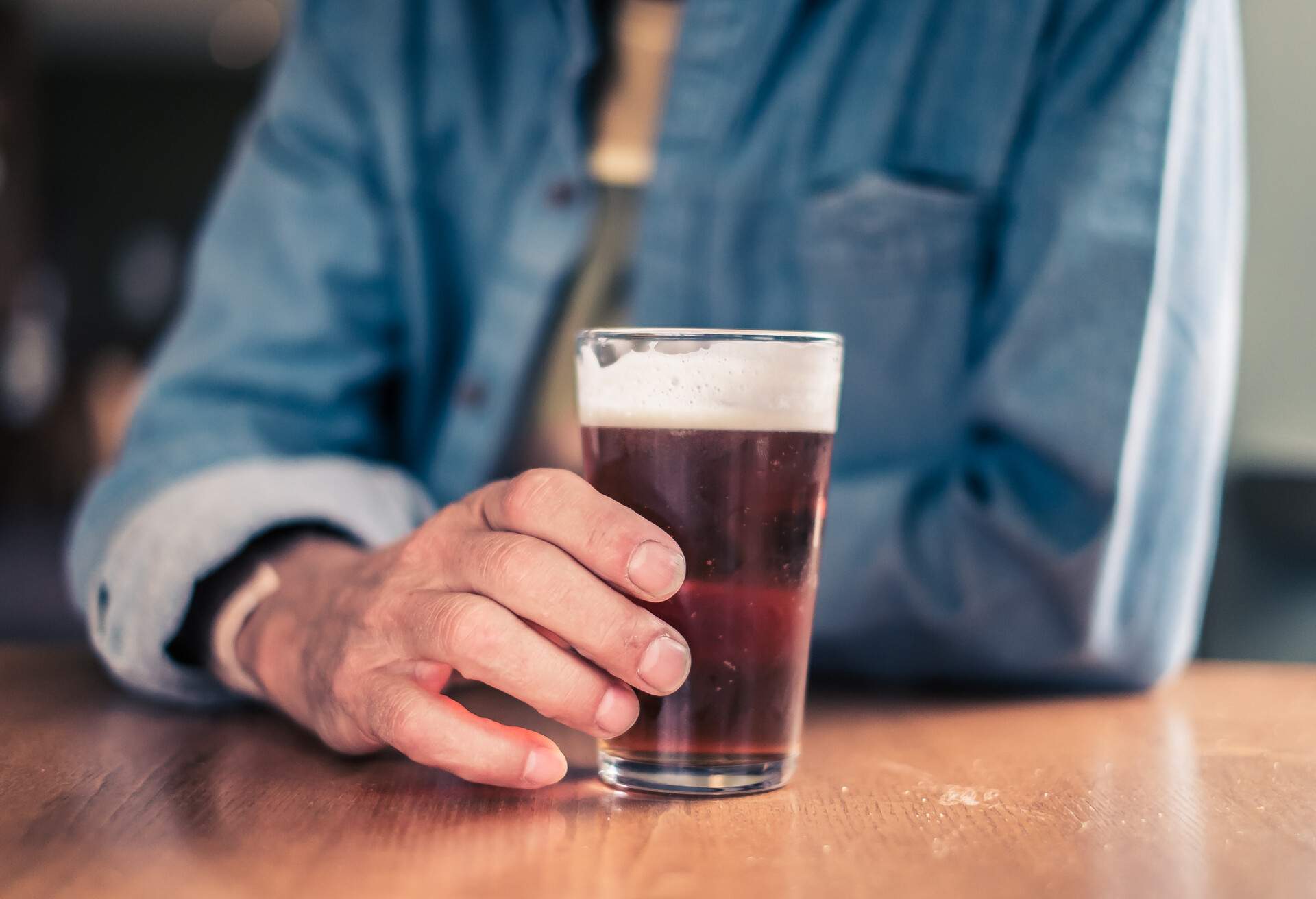 Mid-shot of a man holding an half-pint of beer or real ale, leaning on a table on a Saturday afternoon in an English pub.