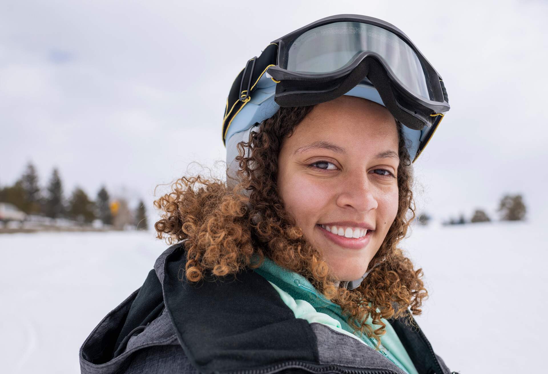 A curly-haired woman smiles while sporting ski goggles and winter clothing.