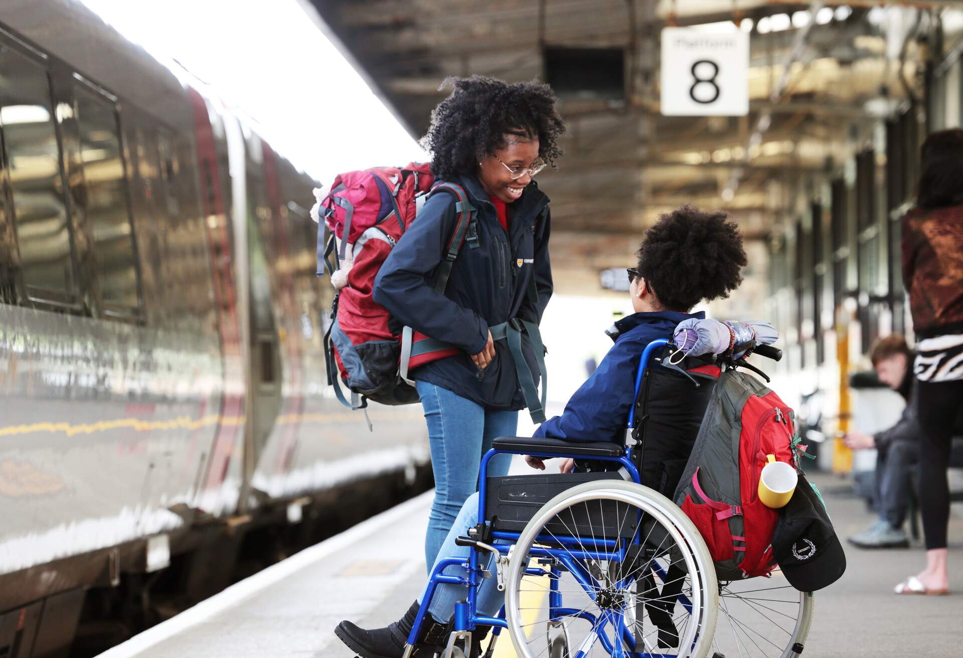 mixed race backpacker disabled young woman in wheelchair talking with friend at station looking at phone