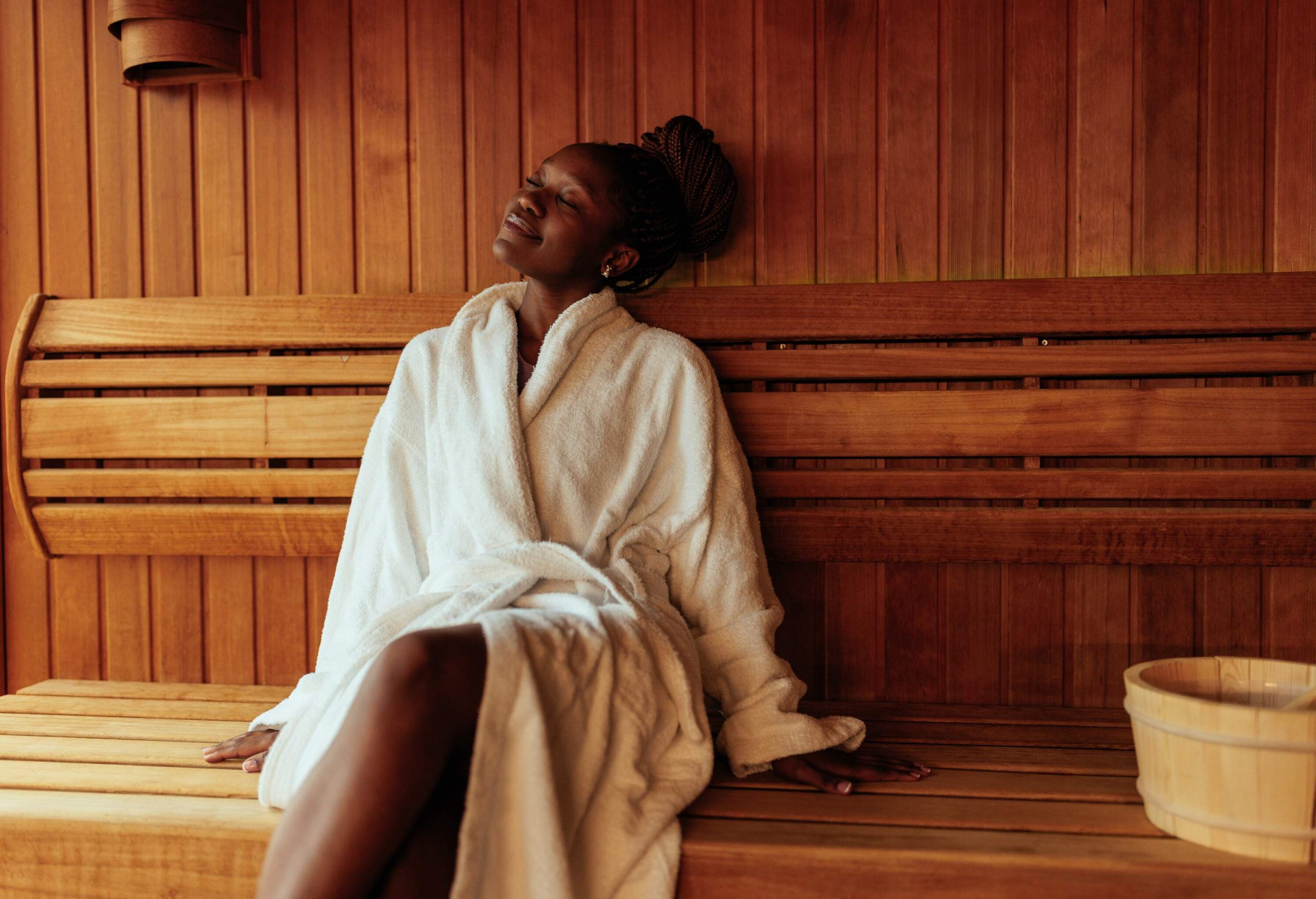 A young black woman is sitting on the bench in the sauna wearing her bathrobe.