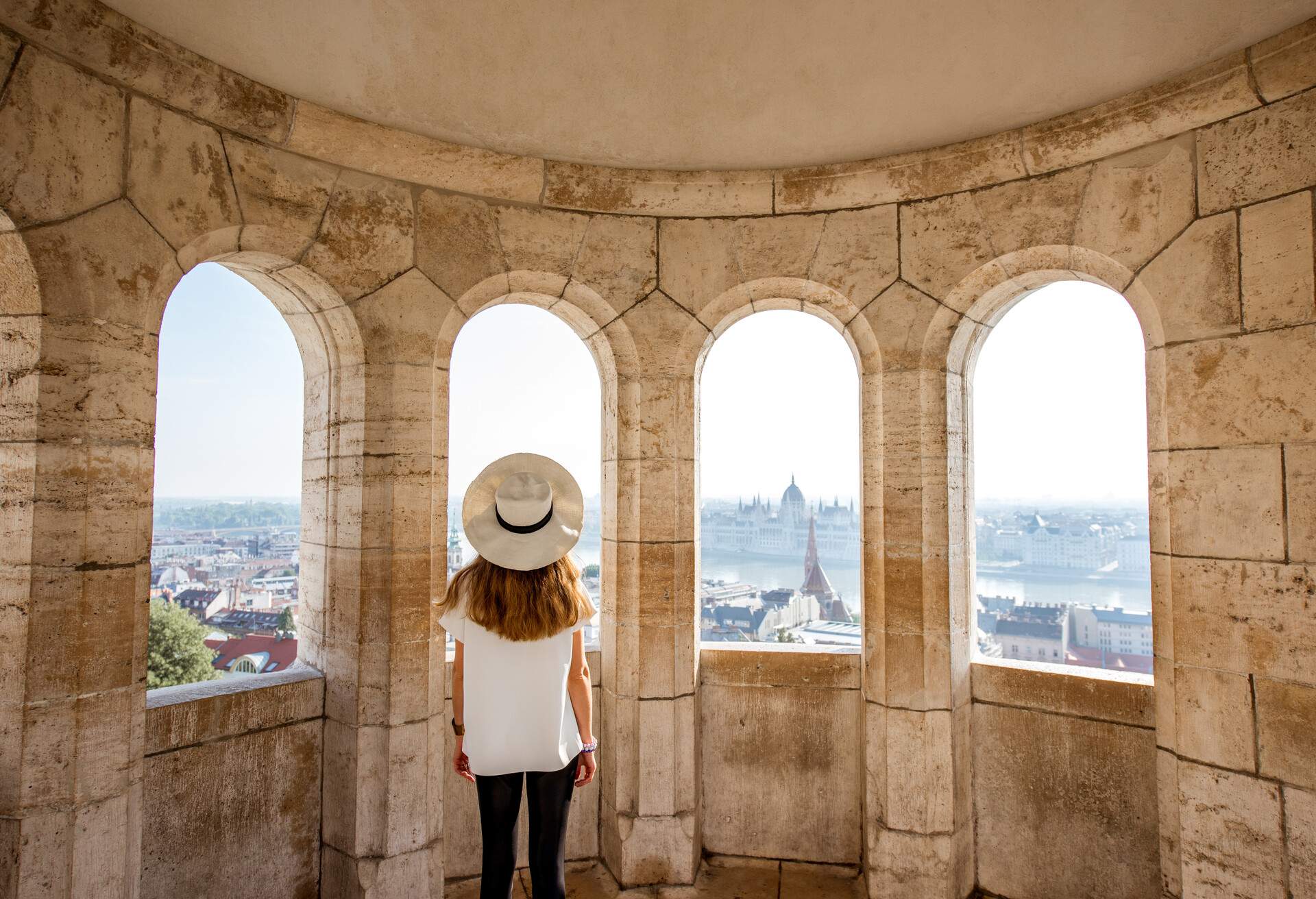 Young woman tourist enjoying cityscape view from the terrace with arches traveling in Budapest, Hungary; Shutterstock ID 1115991713; Purpose: KAYAK MGZN luxury on a shoestring; Brand (KAYAK, Momondo, Any): Any