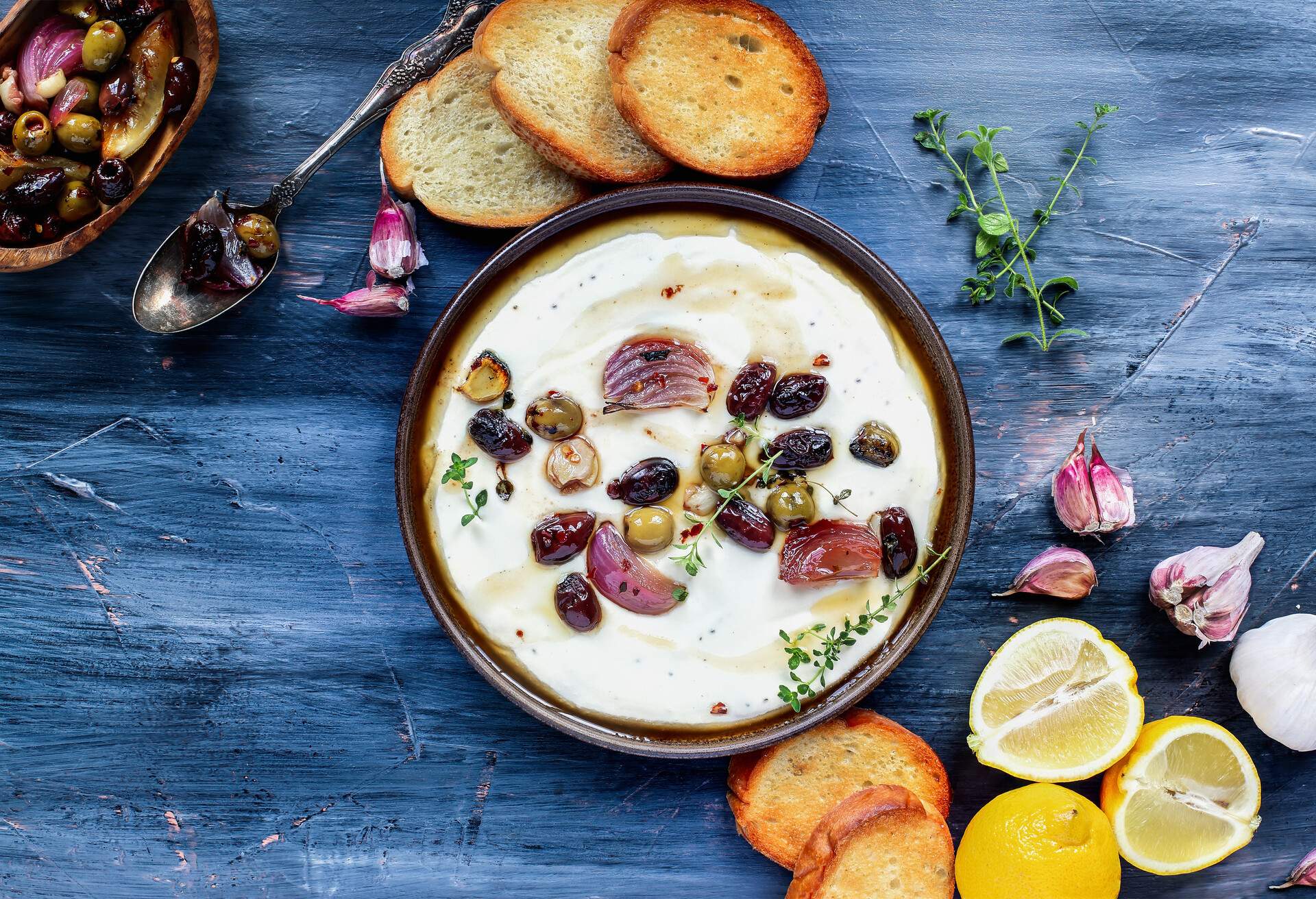 Flatlay of Greek feta cheese dip, Tirokafteri, with roasted olives, Italian garlic, red onions or shallots, drizzled with olive oil and garnished with lemon thyme. Top down view.