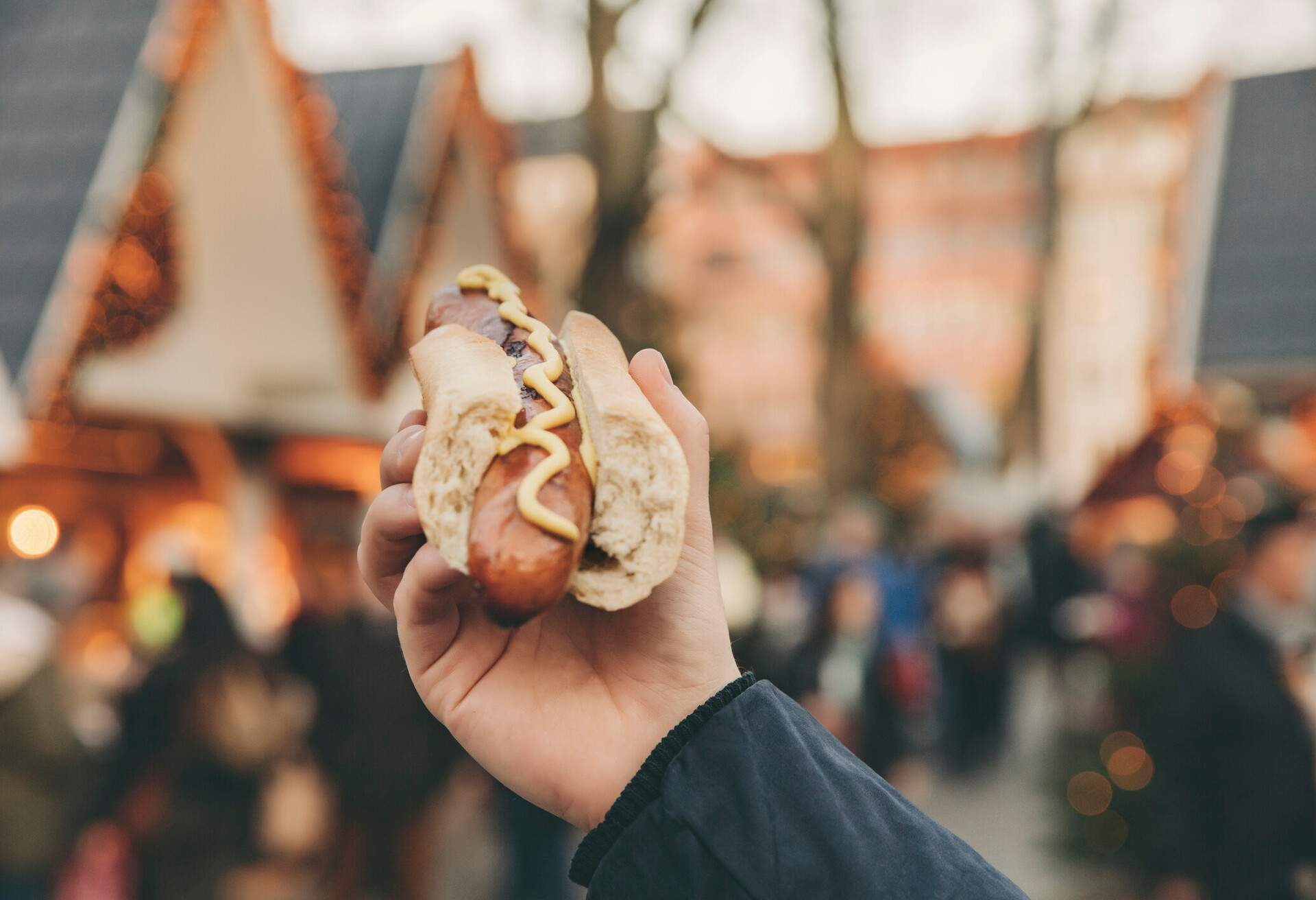 A hand of a person holding a hotdog sandwich on a blurred background of crowded Christmas market.