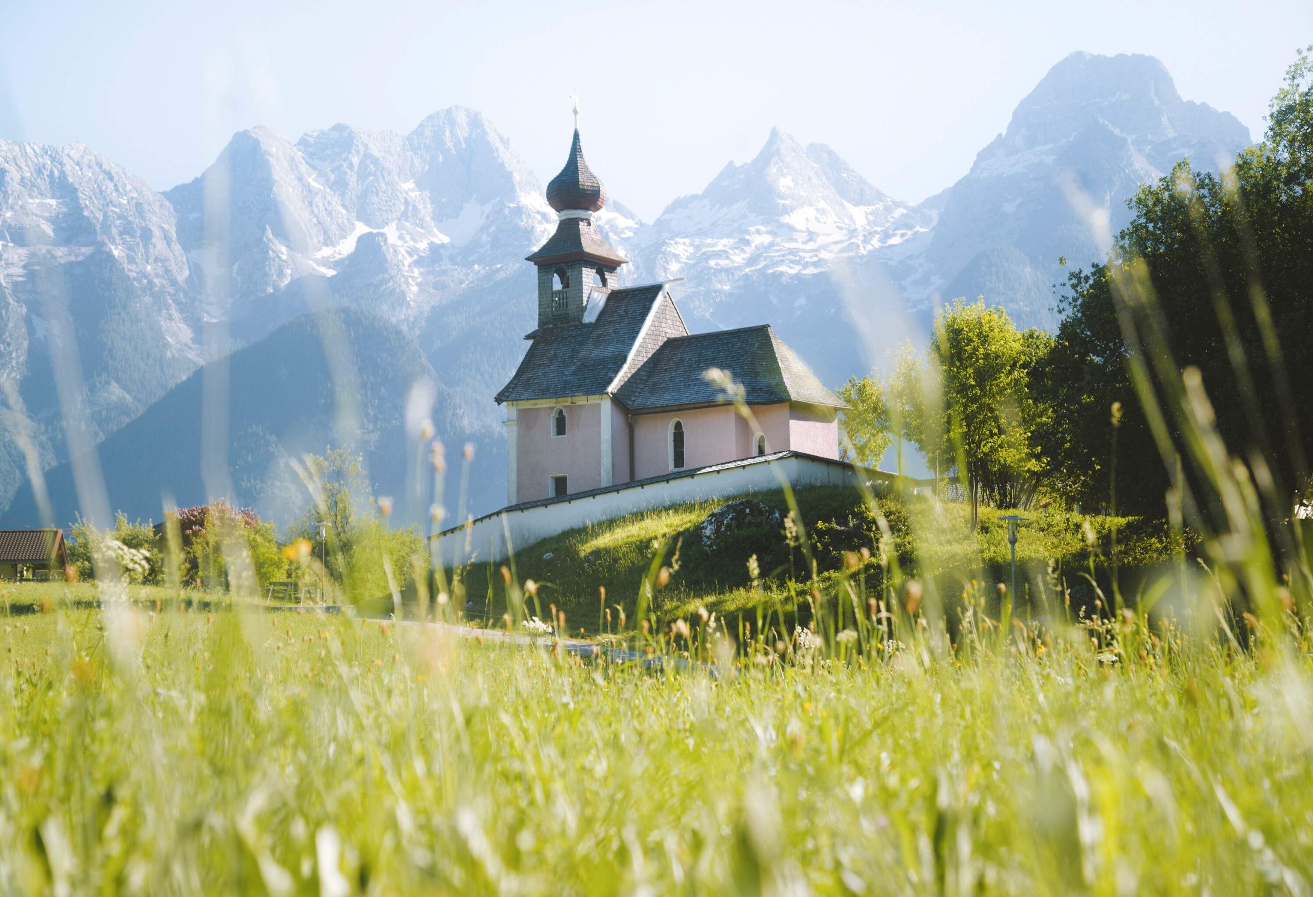 A small and classic chapel on a lush meadow against the rocky mountain range in the background.