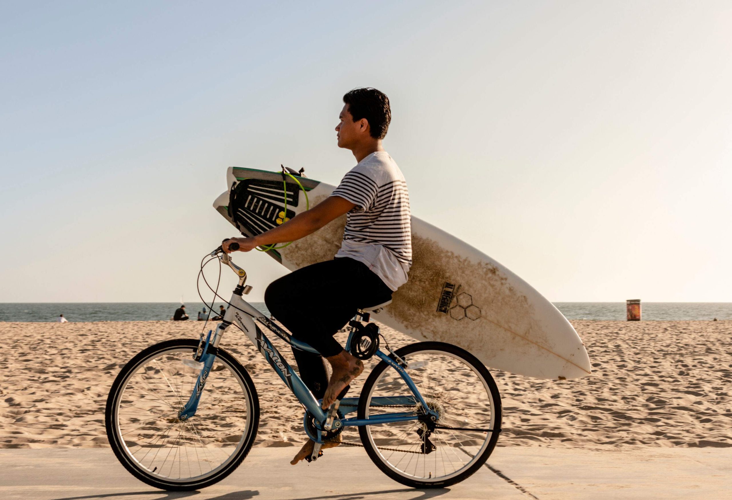 A man riding his bike along the beach with his surfboard gripped to his side.