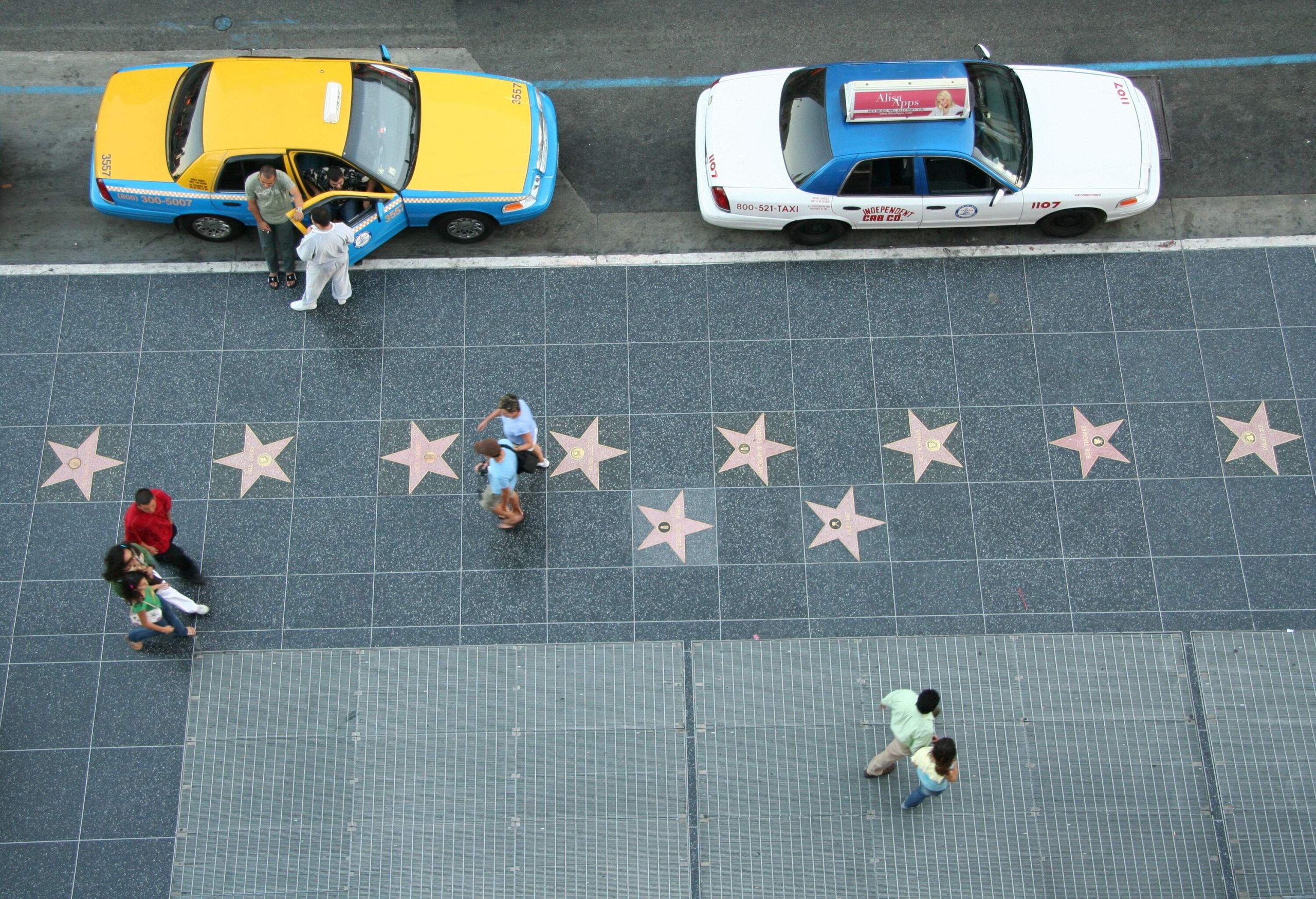 Some people pass by the sidewalk with embedded stars alongside the parked taxicabs.