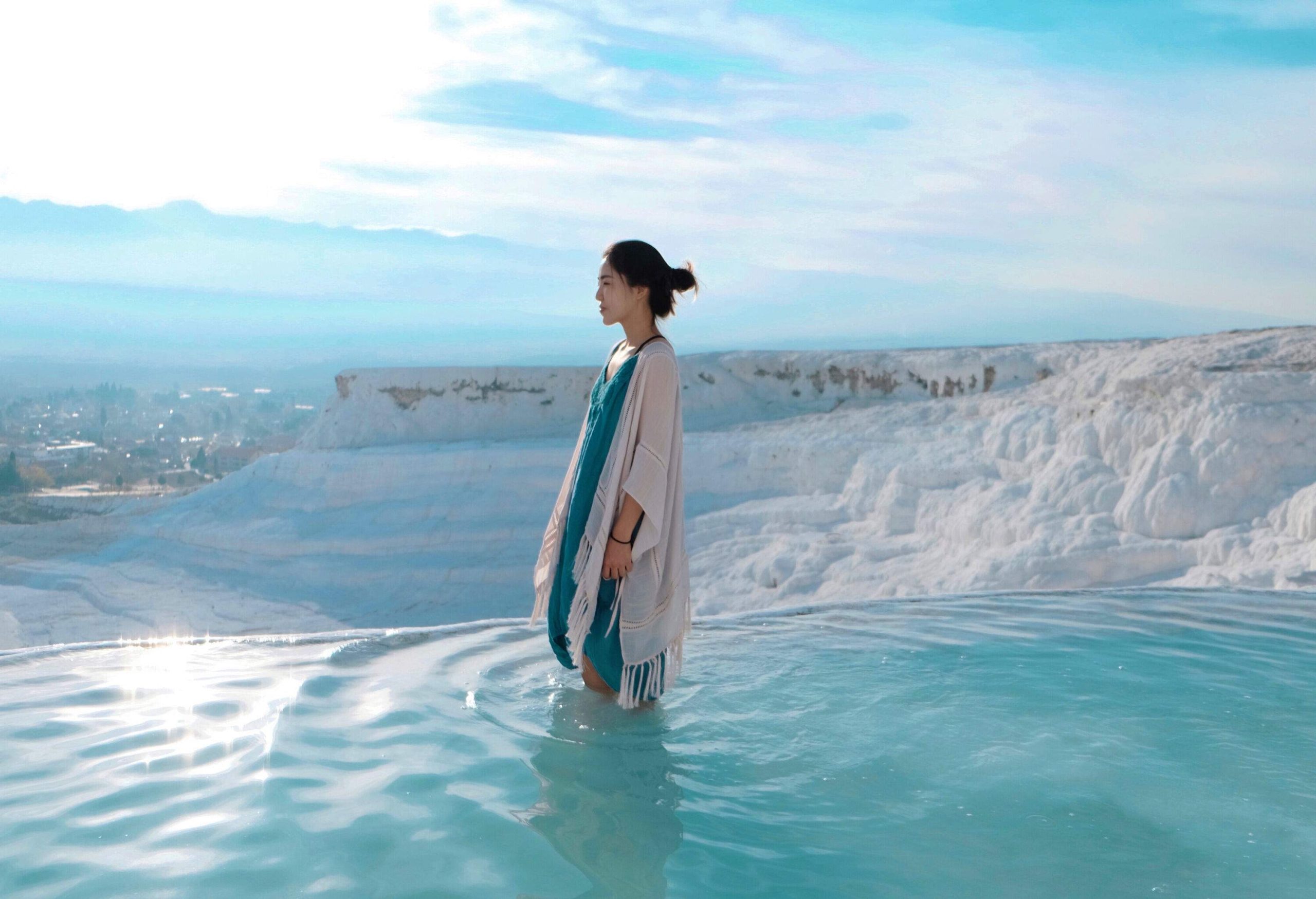 A female stands in a natural turquoise thermal spring pool on a white travertine terrace.