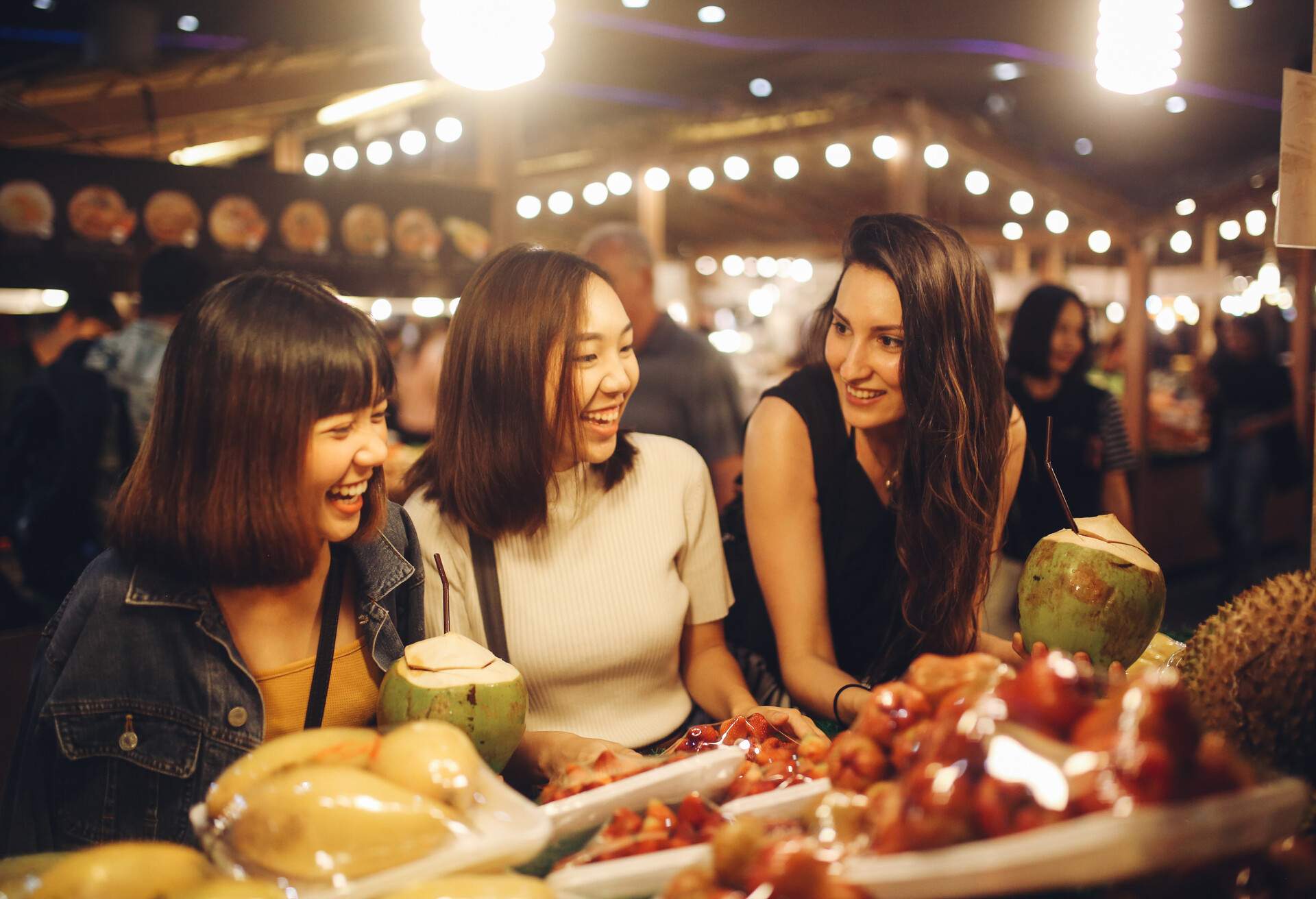 Three women smiling in front of a fruit display on a night market.