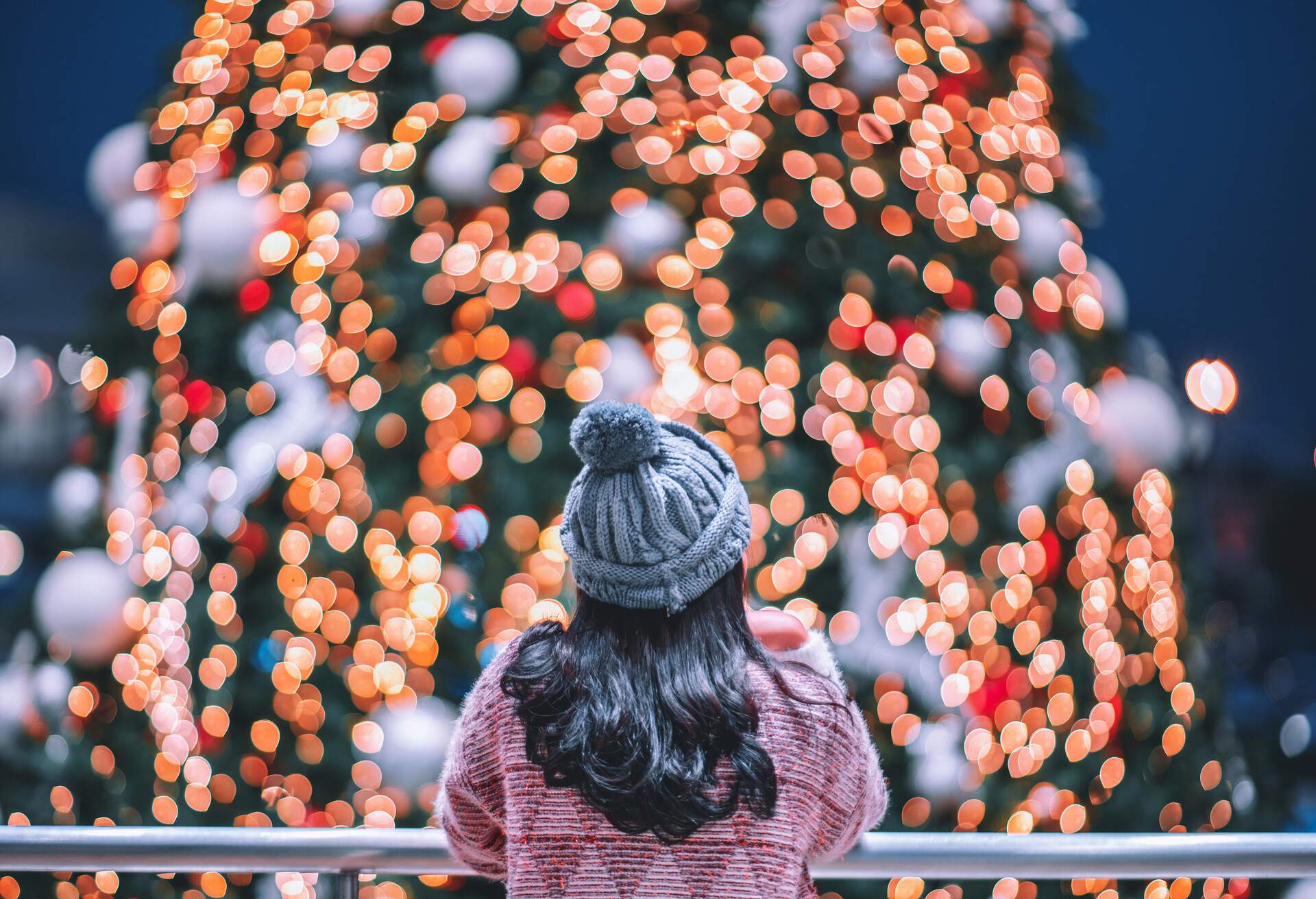 Woman with long hair and a knitted hat leaning on a railing to gaze at a Christmas tree.