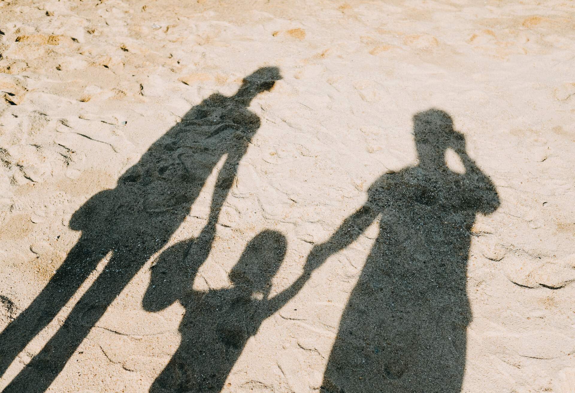 Three people holding hands cast shadows on the sand.