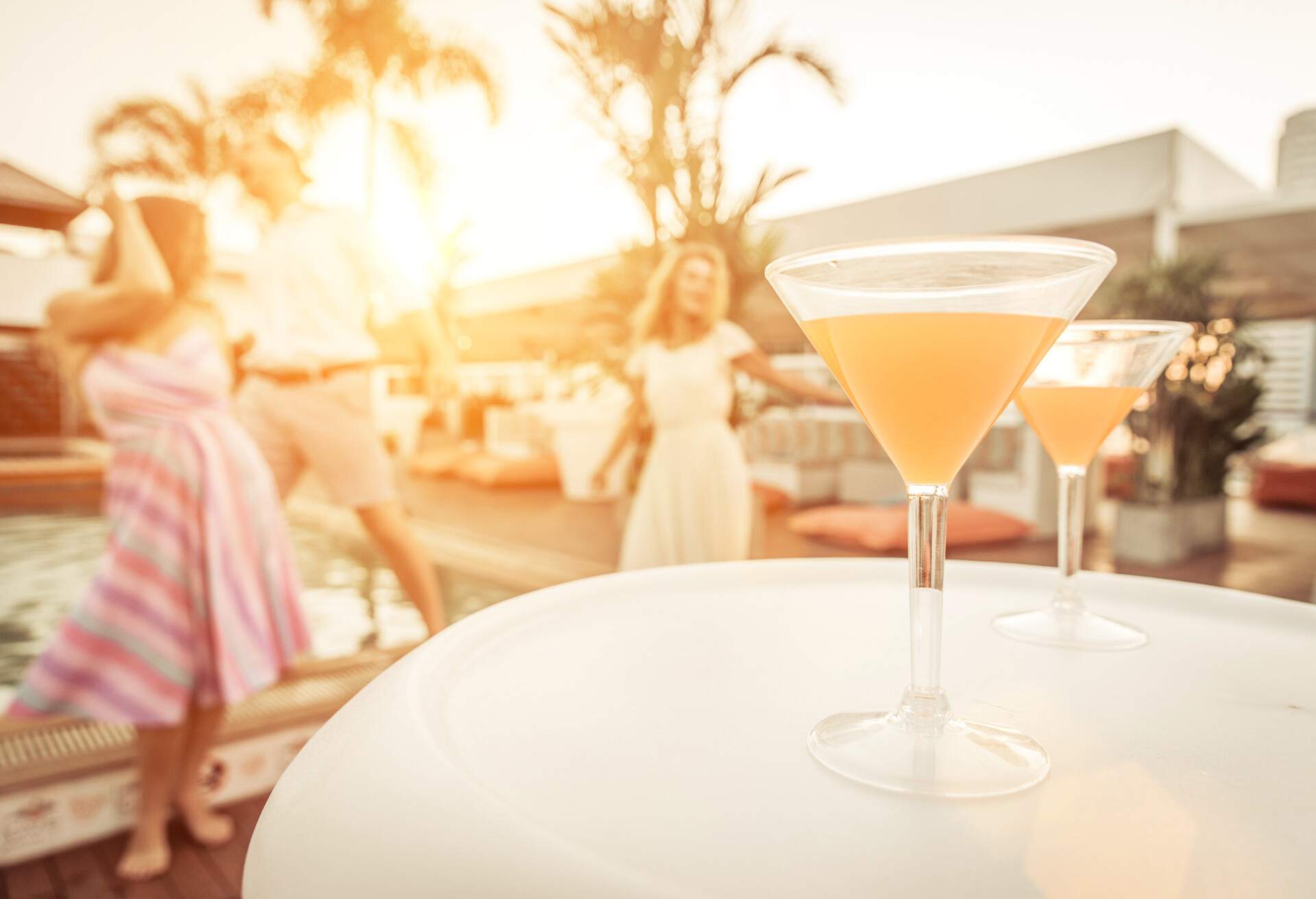 Two yellow cocktail drinks on a white circular table with a background of dancing people.
