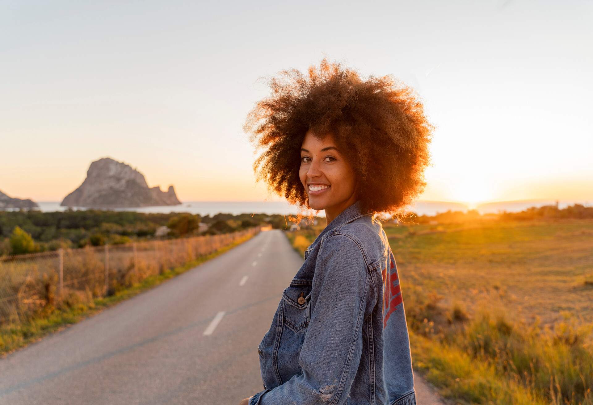A young woman with beautiful afro-textured hair stands on the road in front of a sunset, surrounded by grassy areas, wearing a stylish denim jacket.
