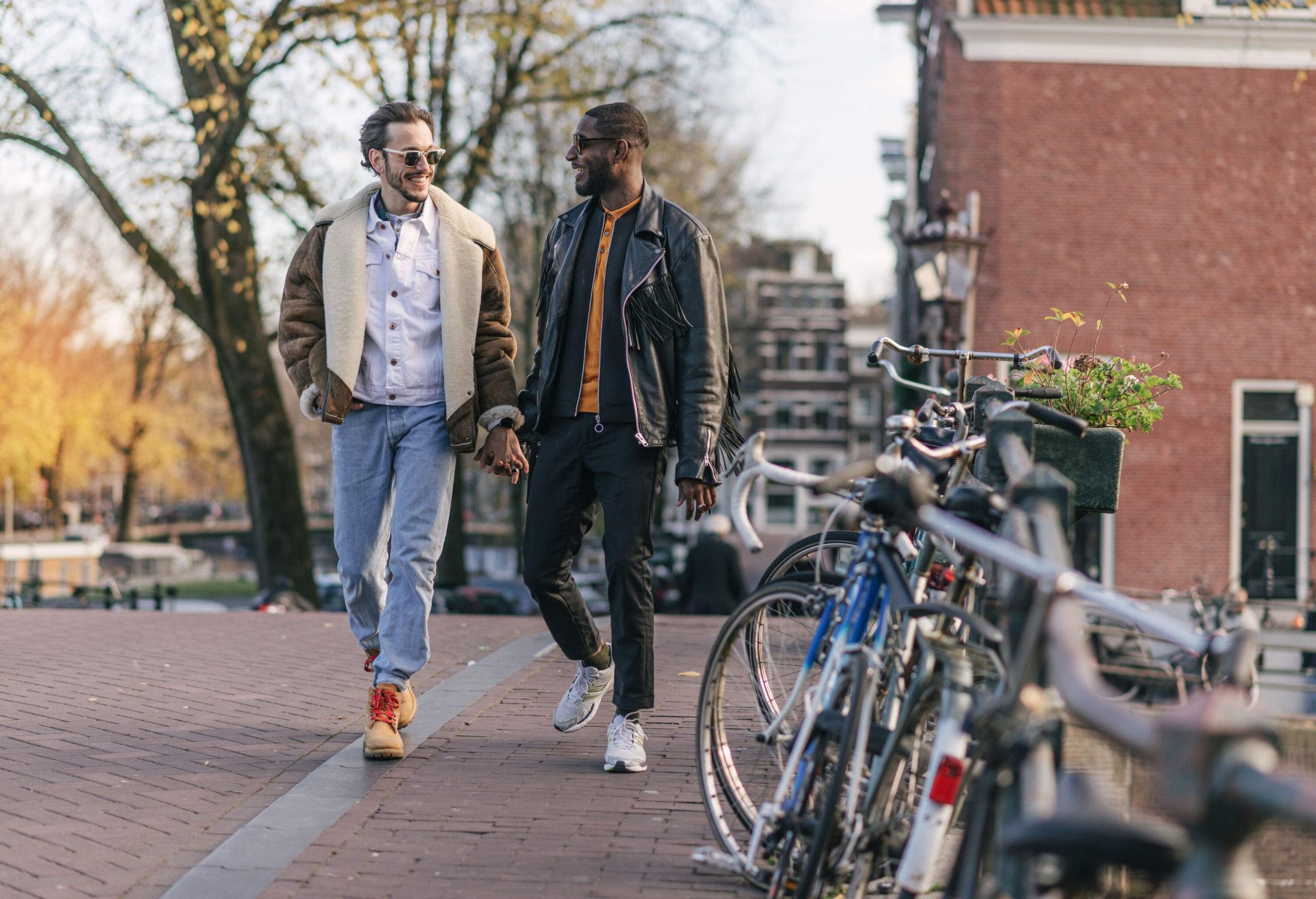 A gay couple in winter clothes walk hand in hand down a cobbled street lined with parked bicycles.