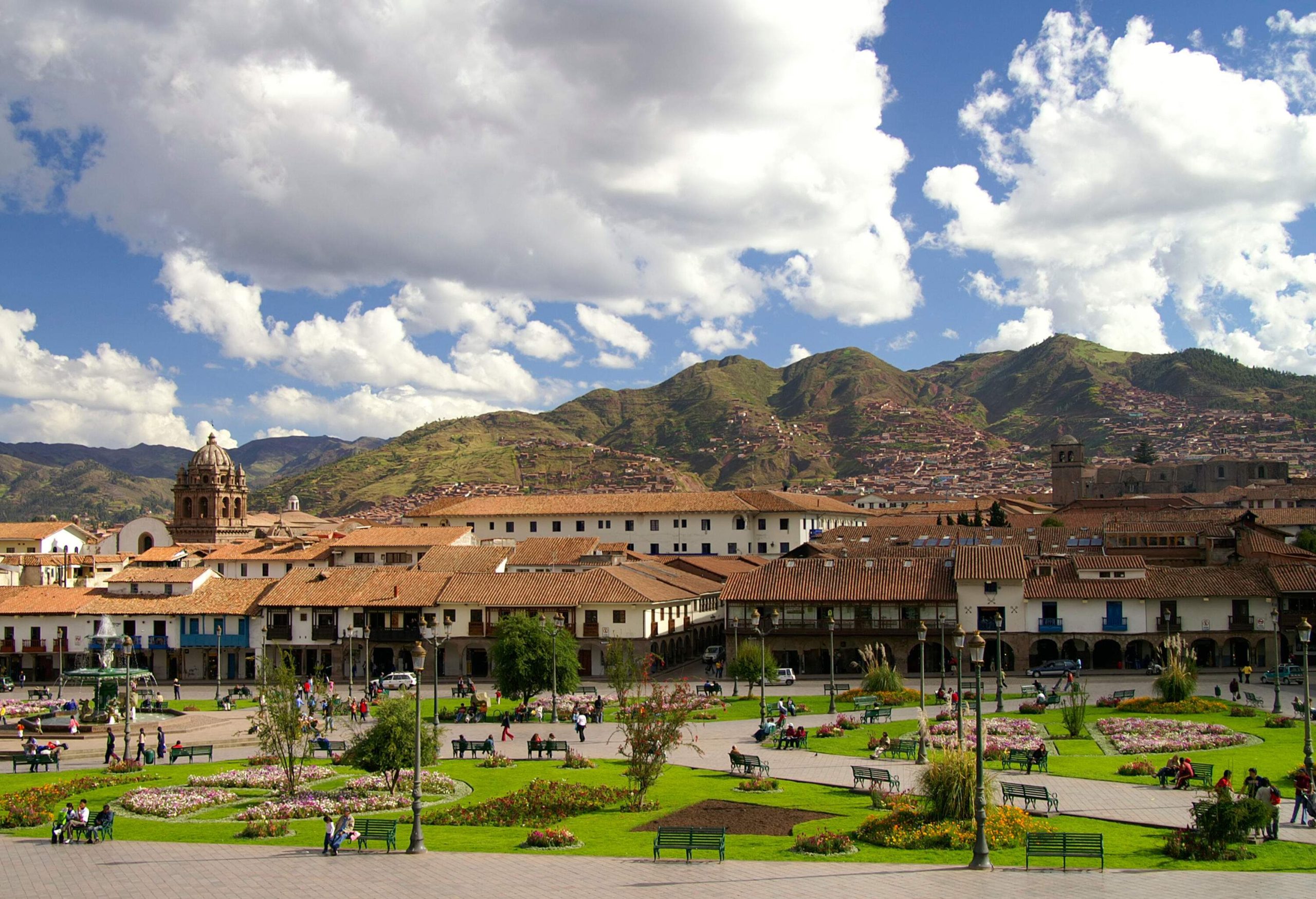 A serene park with its lush green turf adorned with scattered benches and trees stands tall in the background, while buildings stand tall in the background and a majestic mountain range stretches ahead.