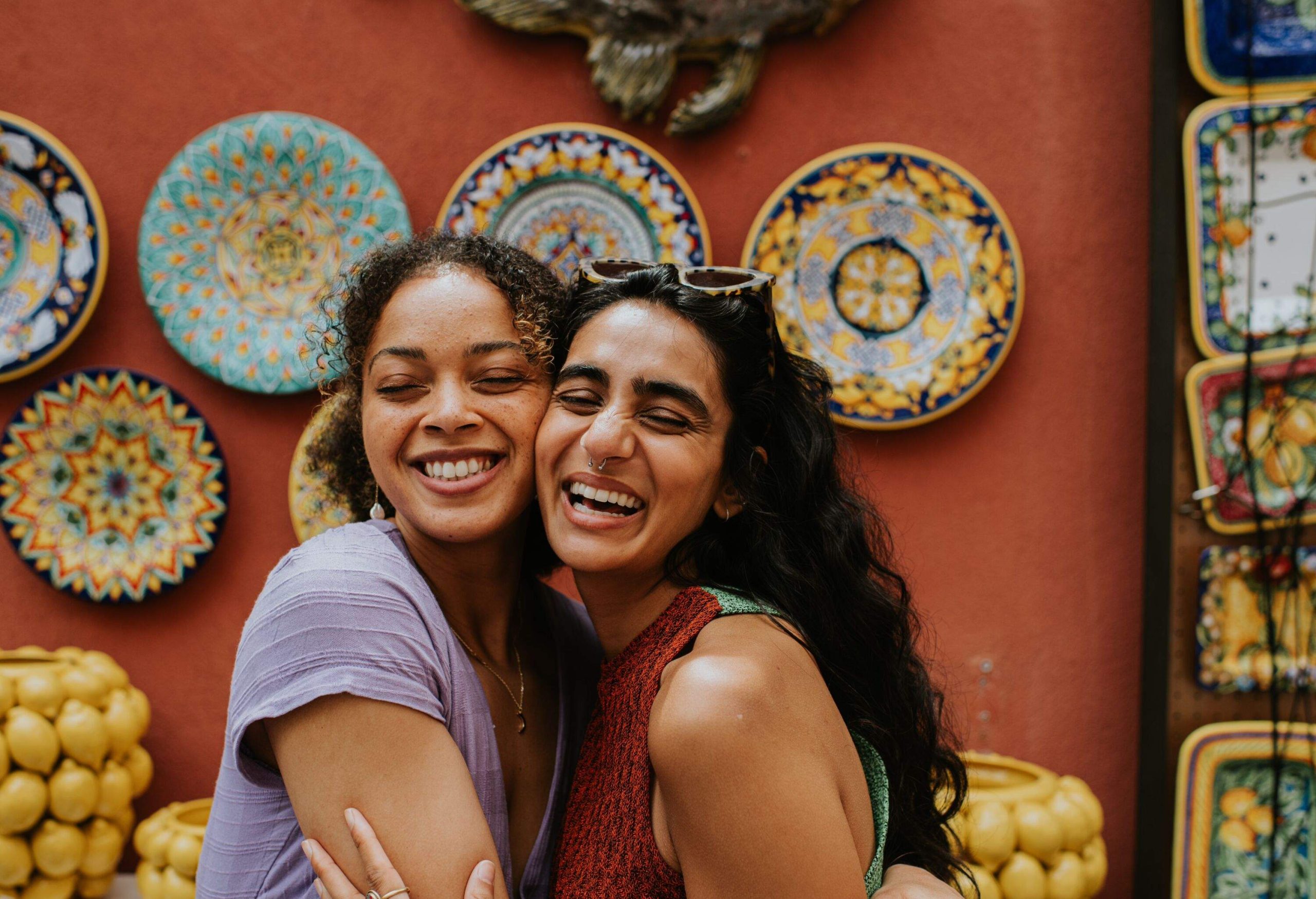 Two young female friends shop in Positano, Italy, famous for its colourful ceramics. They pose together outside a red wall, with wall mounted plates and vases. The woman affectionately embrace as they smile confidently at the camera.