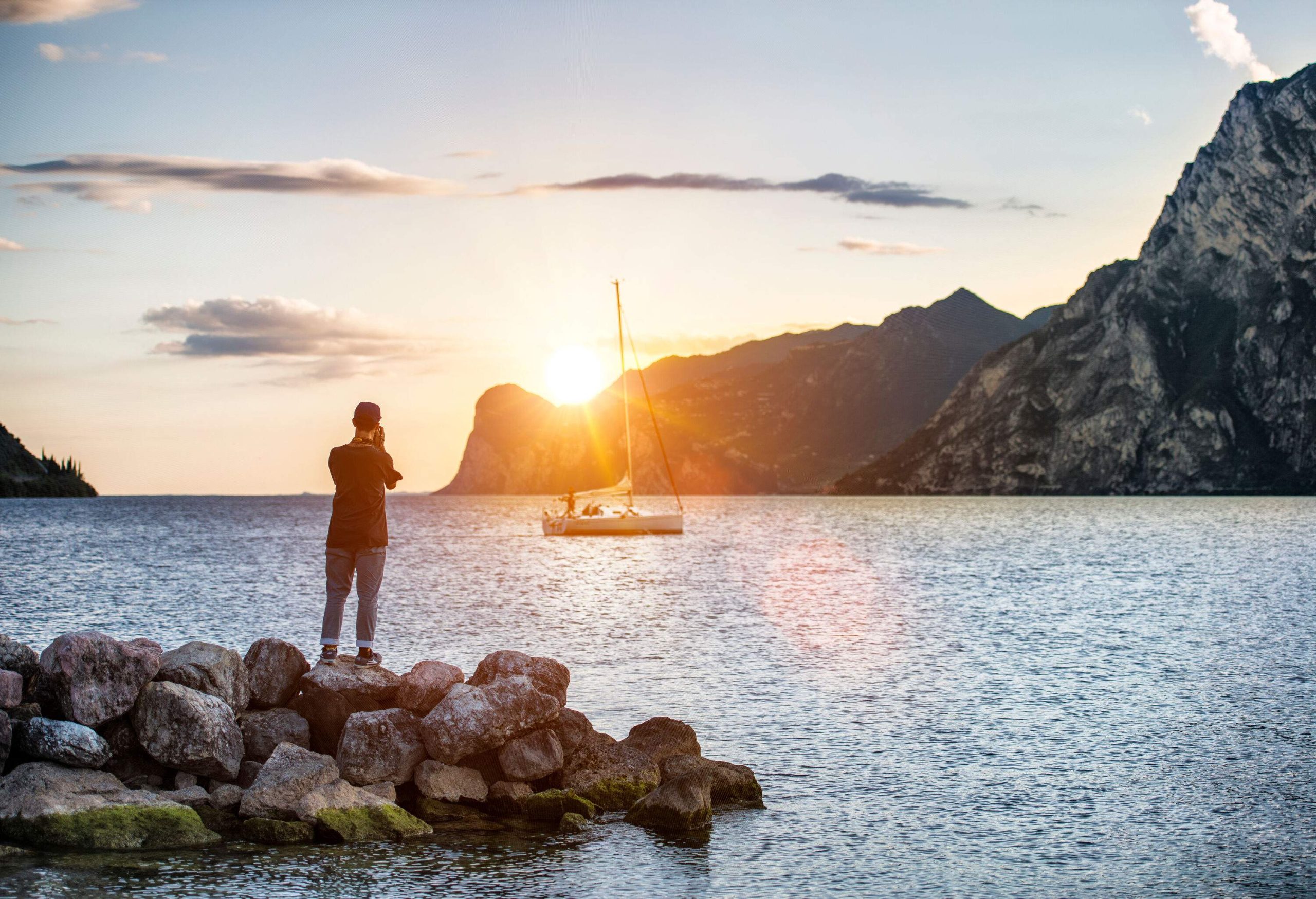 A man standing on a rock taking photos of a sailing boat on a lake surrounded by tall mountains at sunrise.