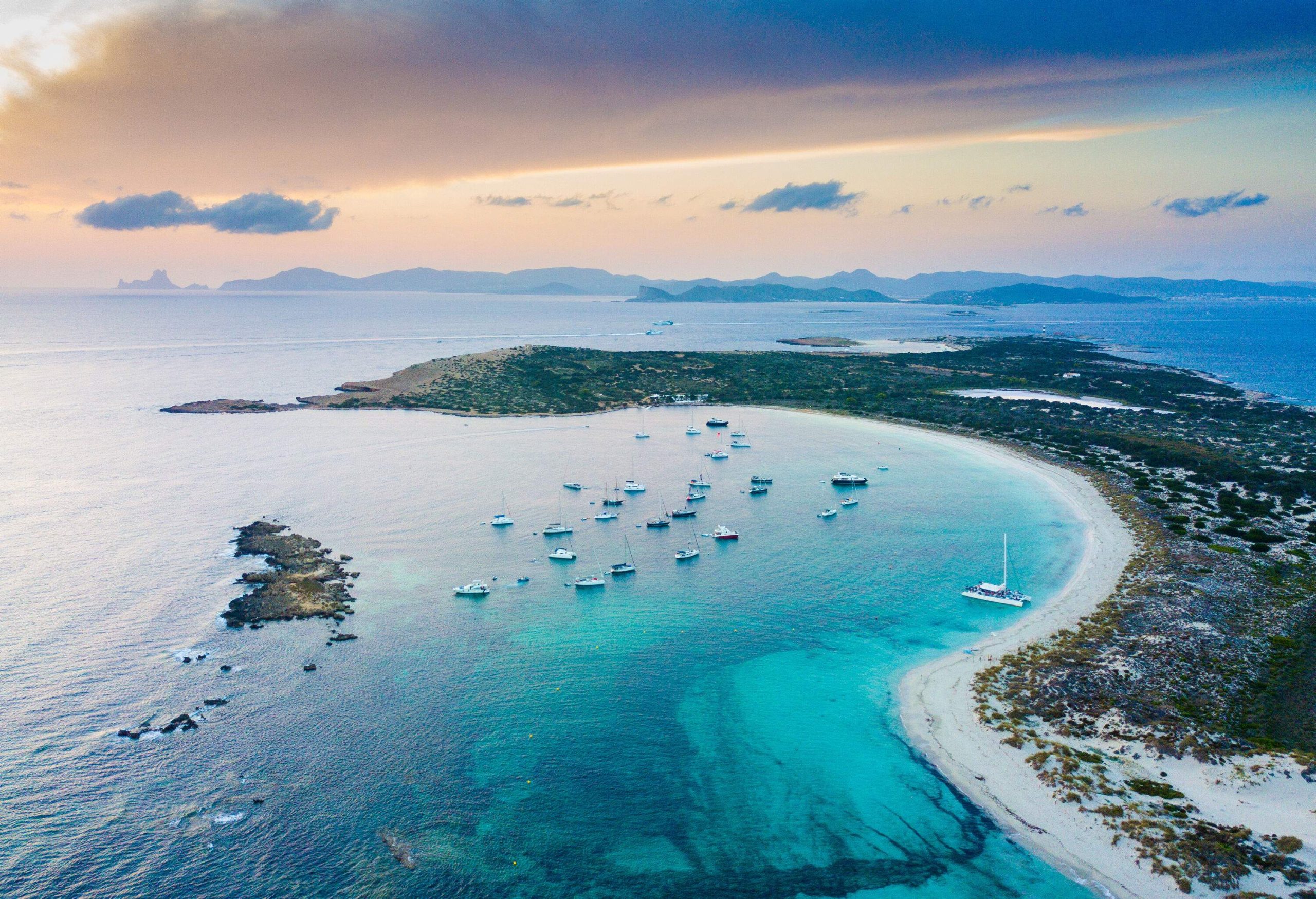 Aerial view of a picturesque beach landscape featuring anchored sailboats and a verdant island against the sunset sky.